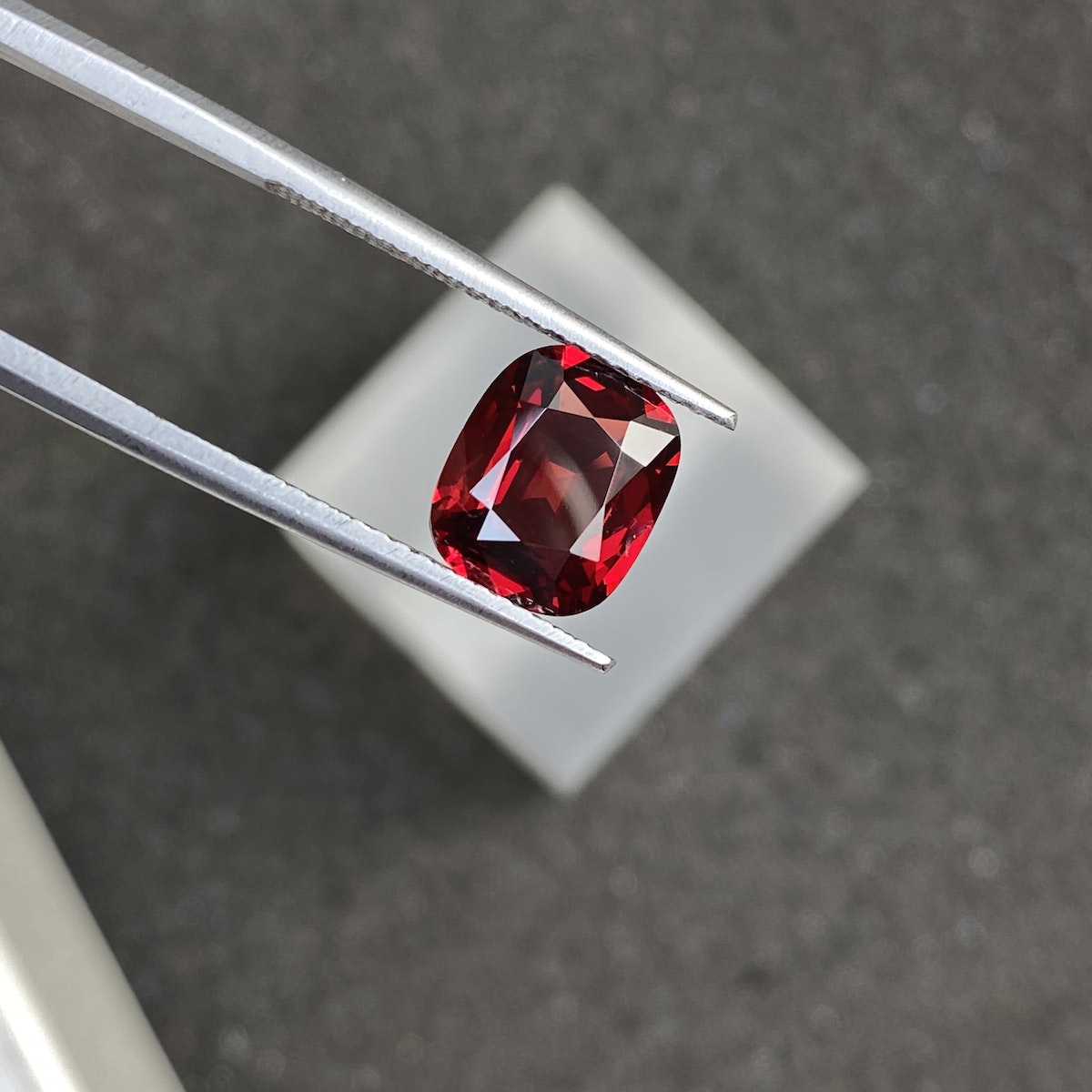 Une paire de pinces maintient le Spinelle Rouge · 2,3 ct de Bonnot Paris sur un fond gris flou, mettant en valeur la clarté de la pierre précieuse taille coussin et sa couleur rouge foncé sous un éclairage focalisé.