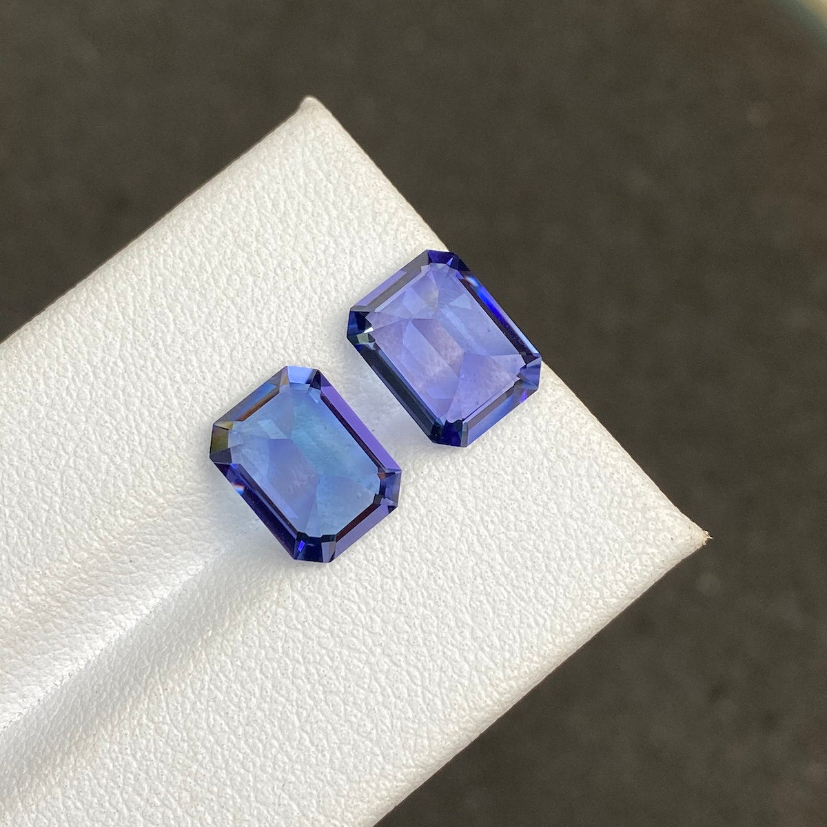 Sur une surface blanche sont présentées deux pierres précieuses Tanzanite à facettes rectangulaires (5,82 ct) de Bonnot Paris, présentant des bords bleu foncé avec des centres plus clairs qui mettent en valeur leur clarté et taillées sur un fond sombre et légèrement flou.