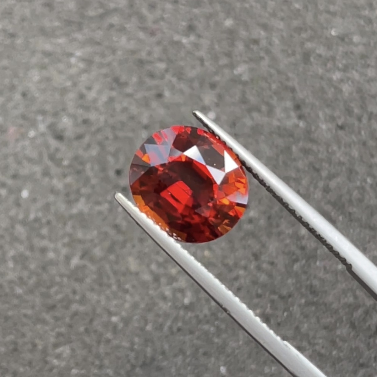 Une pierre précieuse ronde et polie Bonnot Paris Grenat Spessartite · 6,15 ct est serrée dans une pince métallique sur un fond gris texturé, ses facettes rouge foncé reflétant la lumière pour mettre en valeur sa couleur vibrante et sa clarté.