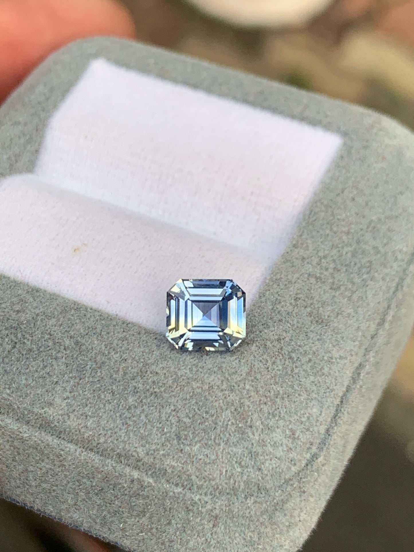 Un Saphir bleu pastel de 2,10 carats de Bonnot Paris, taille émeraude, est présenté dans un écrin en velours gris. Cette pierre précieuse sri-lankaise présente une surface réfléchissante aux facettes aux nuances bleues envoûtantes.