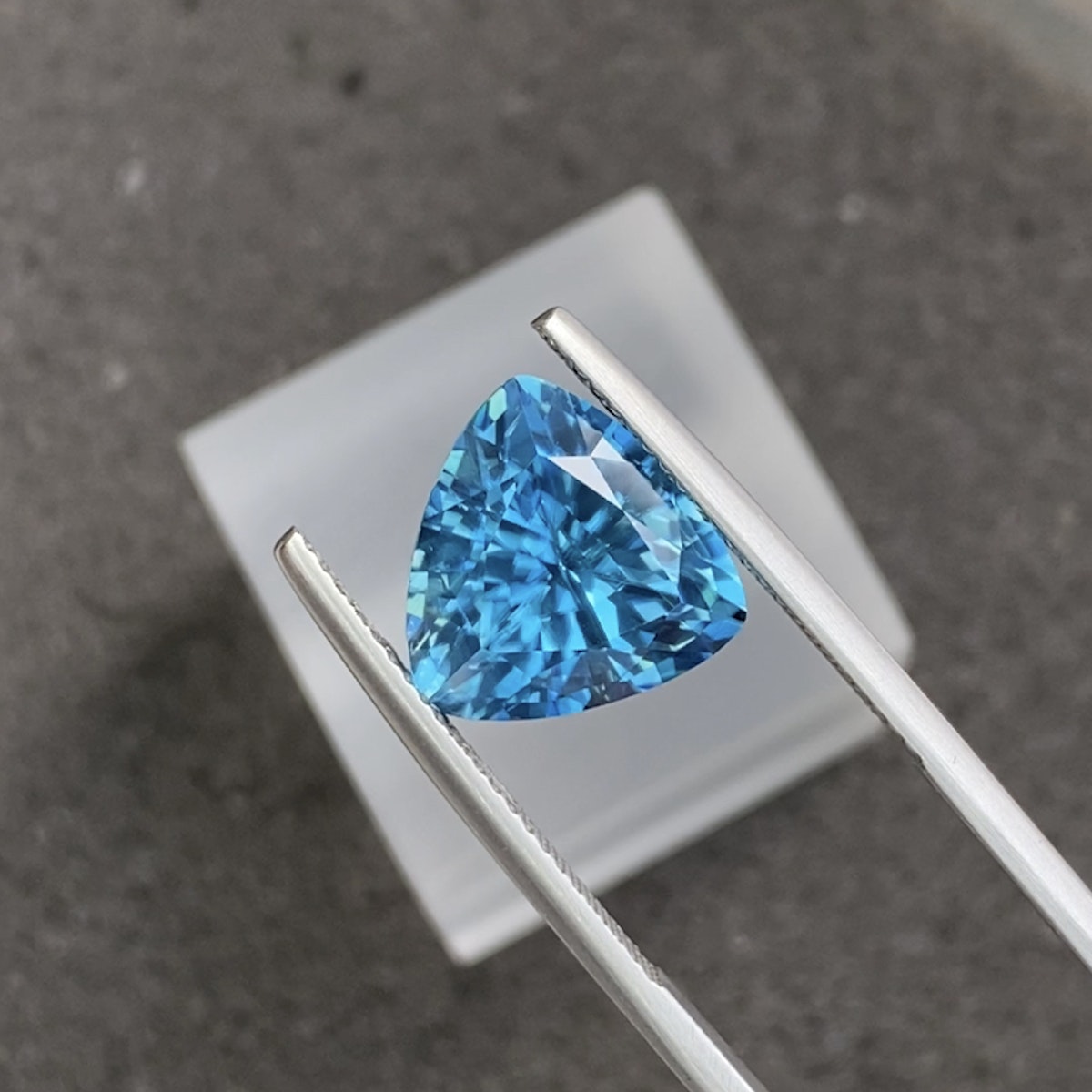 Une pince à épiler tient le Zircon Bleu taille trilliant · 7,96 carats de Bonnot Paris sur un fond gris. Ses nombreuses facettes captent et reflètent la lumière pour sublimer sa couleur vibrante. Une surface carrée blanche est partiellement visible à l'arrière-plan.