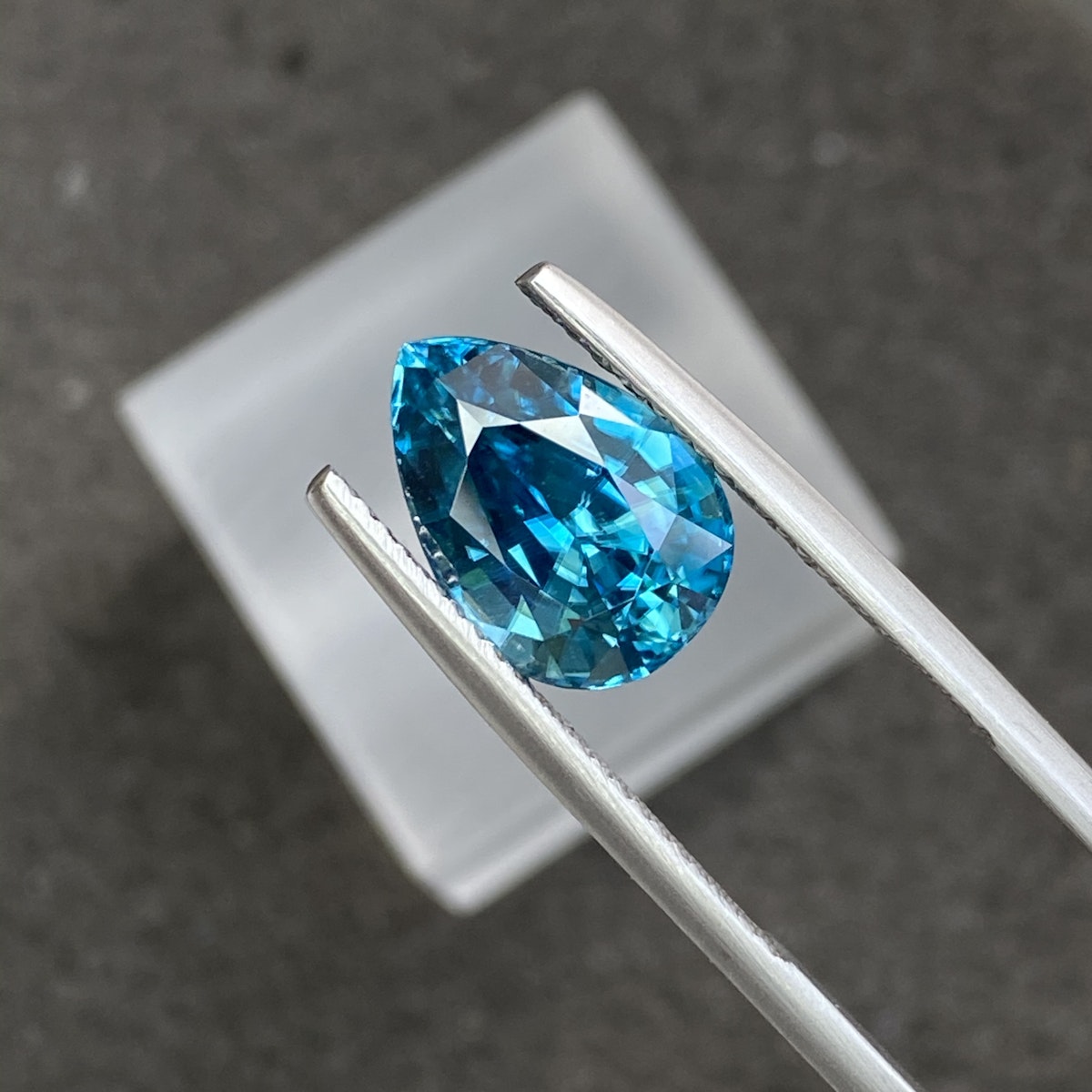 Une pierre précieuse bleue en forme de poire, le Zircon Bleu · 6,72 carats de Bonnot Paris, est maintenue entre des pinces en argent sur un fond sombre, avec un carré blanc flou en arrière-plan. La pierre scintille, révélant des facettes qui reflètent la lumière.