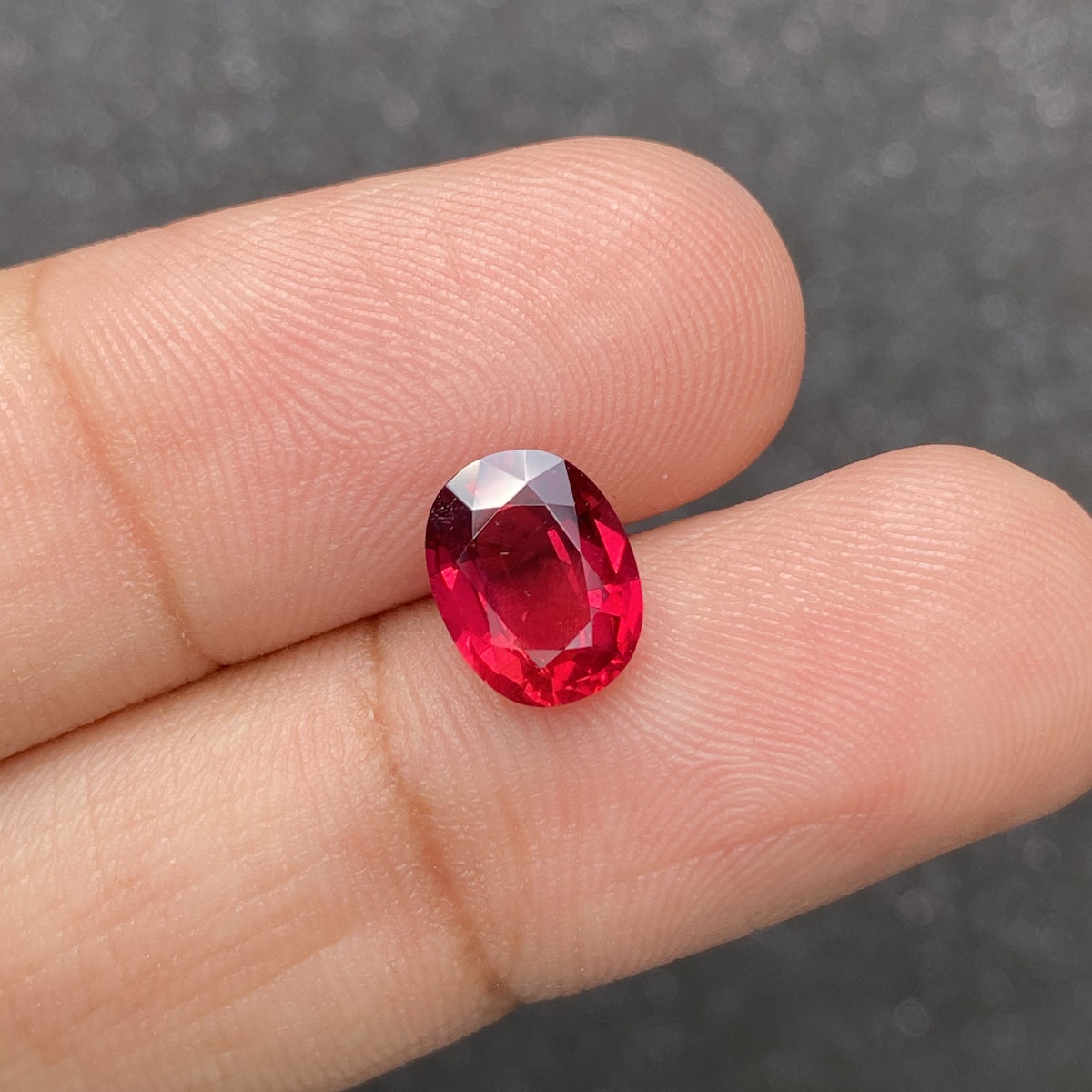 Gros plan d'une main tenant le Rubis du Mozambique de Bonnot Paris, une pierre précieuse ovale facettée rouge de 2,59 carats. Un fond gris foncé flou met en valeur la couleur vibrante de la pierre.