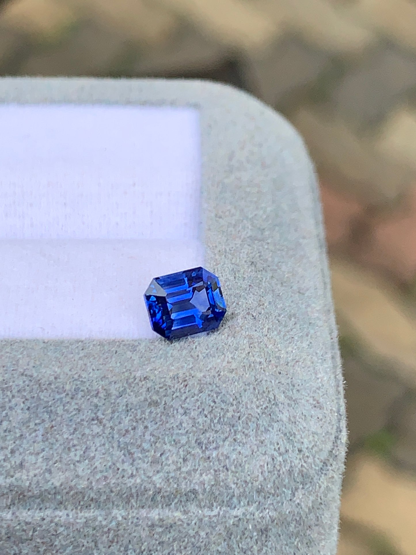 Le Saphir bleu • 1,03ct de Bonnot Paris est élégamment serti sur un écrin gris clair ; ses facettes taille émeraude captent et reflètent magnifiquement la lumière sur un fond doux et terreux.
