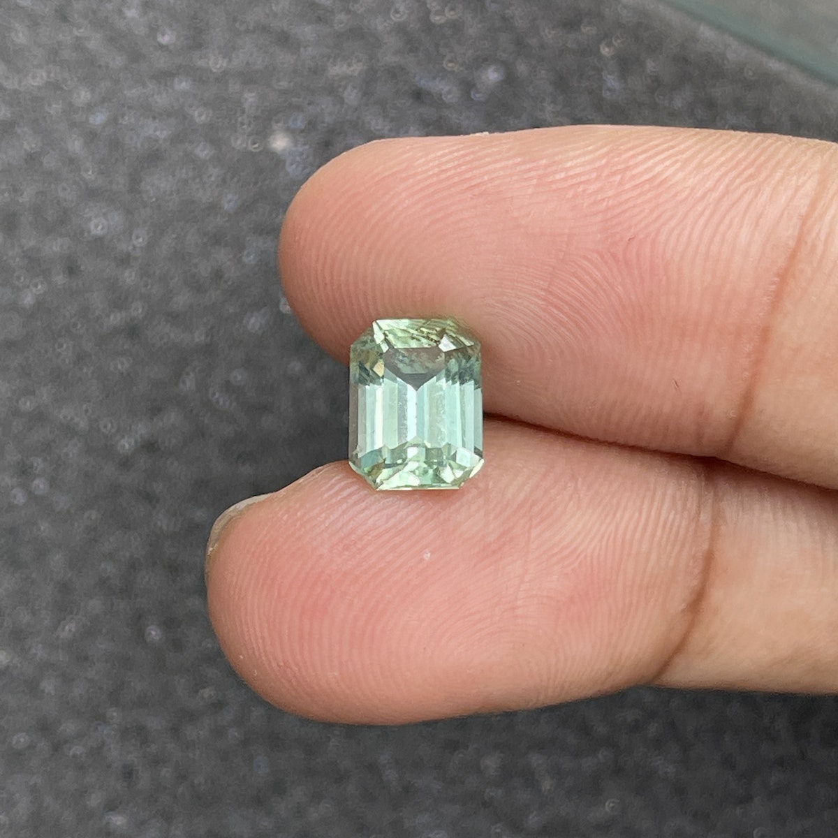Gros plan d'une main tenant la Tourmaline Verte Bonnot Paris · 2,43 ct, une pierre précieuse vert clair taille émeraude, entre deux doigts sur un fond gris texturé.