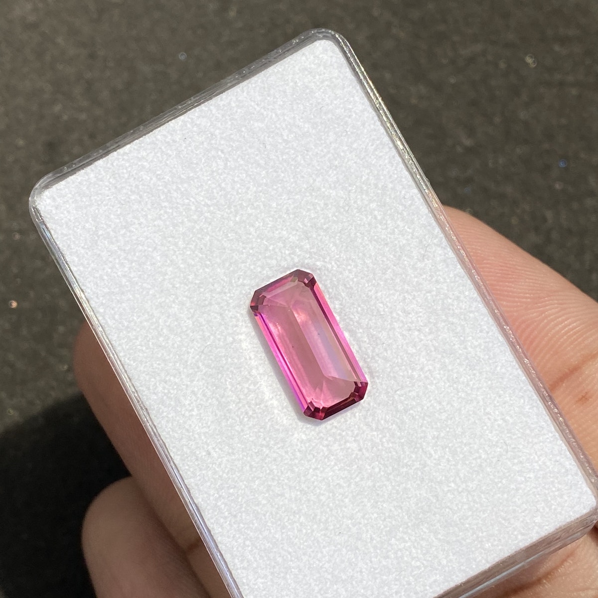 Une Tourmaline Rose de 3,95 carats de Bonnot Paris, vibrante et claire, est présentée alors que les doigts la tiennent contre une carte blanche, mettant en valeur sa pierre précieuse rose rectangulaire facettée qui reflète magnifiquement la lumière.