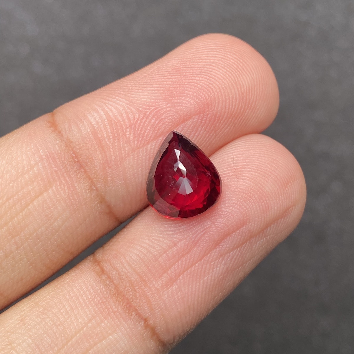 Une main tient le Rubis du Mozambique · 4,03 ct de Bonnot Paris, une petite pierre précieuse rouge en forme de larme avec une surface facettée, sur un fond gris texturé.