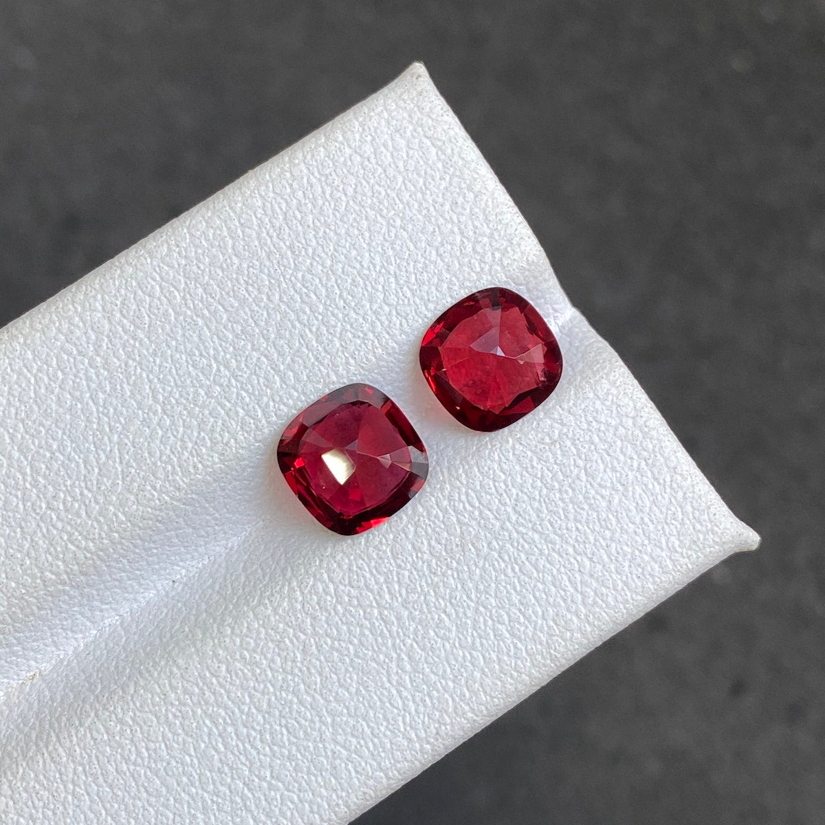 Deux spinelles rouges taille coussin de Bonnot Paris, chacune de 3,49 carats, sont présentées sur une surface blanche sur fond sombre. Elles présentent une couleur rouge intense et profonde, avec des facettes qui reflètent brillamment la lumière.