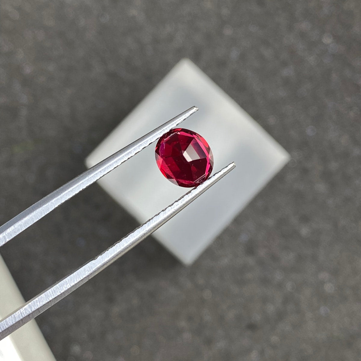 Une paire de pinces métalliques maintient la pierre précieuse Spinelle Rouge · 1,25 ct de Bonnot Paris, une pierre précieuse rouge foncé de forme ovale, sur un fond gris flou avec un carré blanc en dessous.