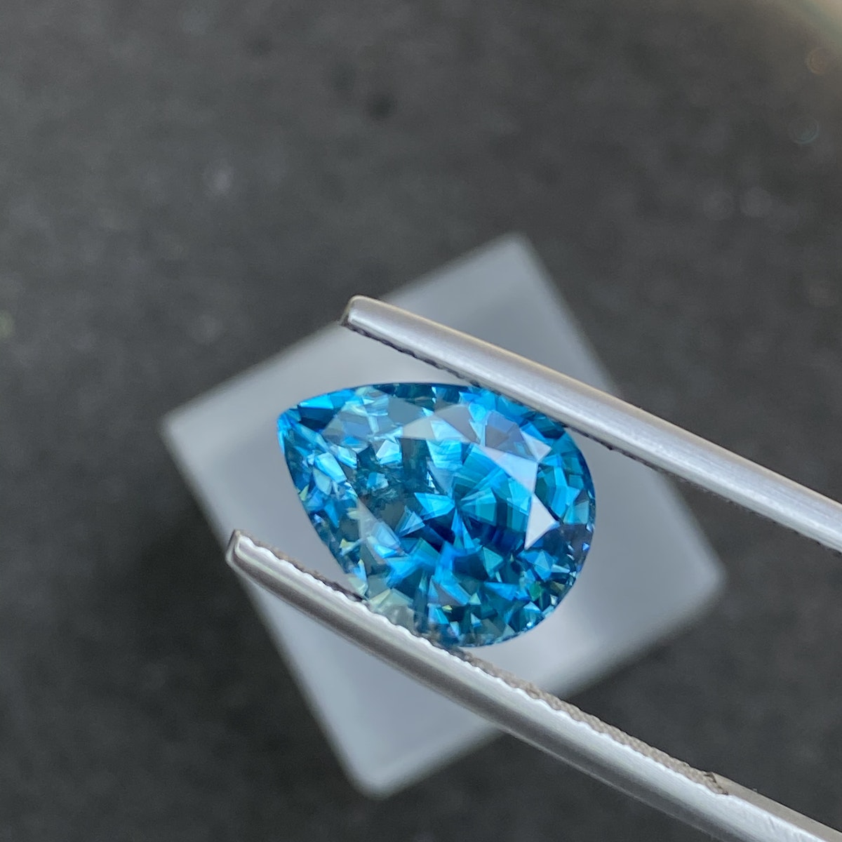 Un Zircon Bleu de 7,75 carats de Bonnot Paris, en forme de larme et maintenu par une pince à épiler sur un fond sombre flou, présente un éclat facetté brillant.