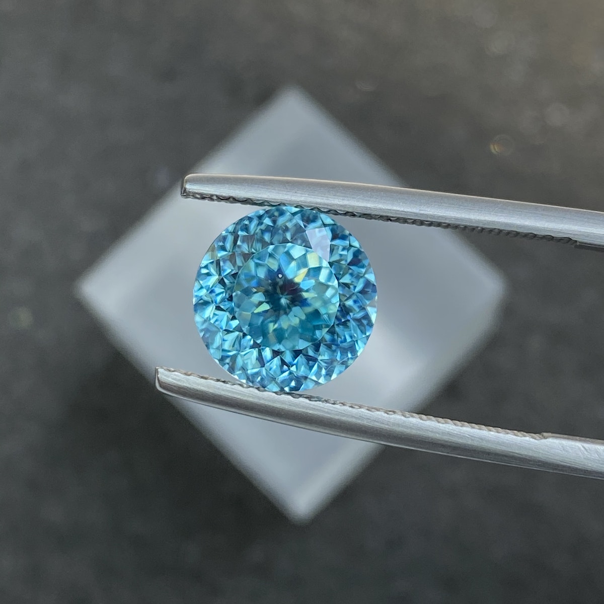 Une pince en argent maintient le Zircon Bleu · 7,56ct de Bonnot Paris sur un fond sombre flou, avec ses facettes bleues scintillant à la lumière.
