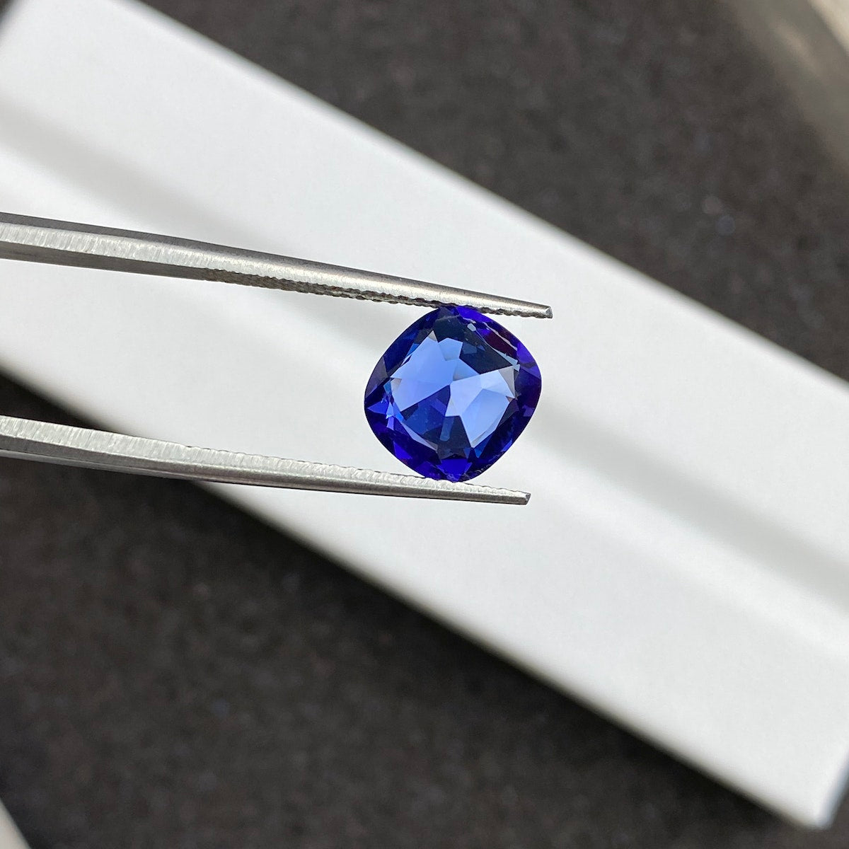 Gros plan sur la tanzanite Bonnot Paris de 2,44 carats, une pierre précieuse bleue taille coussin, maintenue par une pince métallique sur un fond flou. Ses facettes reflètent la lumière, mettant en valeur sa teinte bleu profond.