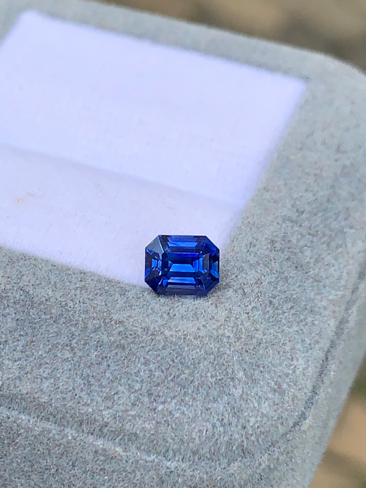 Un Saphir bleu taille émeraude de 1,03 ct, d'un bleu éclatant, signé Bonnot Paris, repose sur une surface coussinée gris clair inclinée. Cette pierre précieuse scintille à la lumière, sa couleur riche et ses facettes savamment taillées mettant en valeur un bijou exquis et unique.