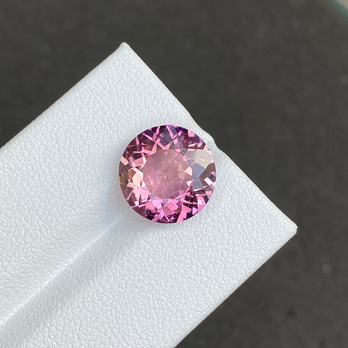 La Tourmaline Cuprifère · 5,22 carats de Bonnot Paris est mise en valeur sur une surface blanche texturée. Ses tailles rondes et facettées reflètent la lumière, soulignant ses teintes roses vibrantes sur un fond sombre, sublimant ainsi son éclat.