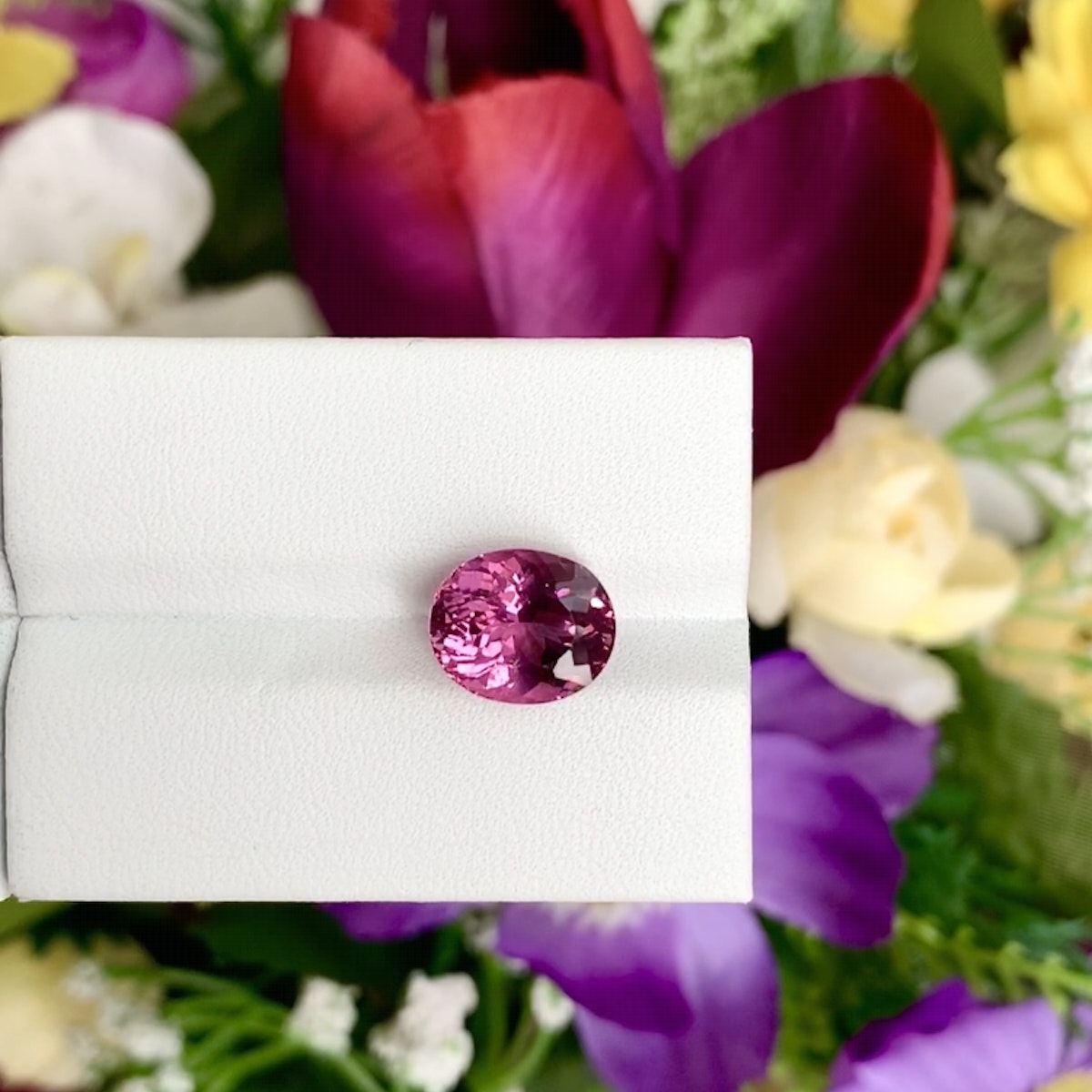 Une Tourmaline Rubellite ronde facettée (4,35 ct) de Bonnot Paris est présentée sur un support blanc avec un fond floral vibrant et flou dans des teintes violettes, jaunes, blanches et vertes.