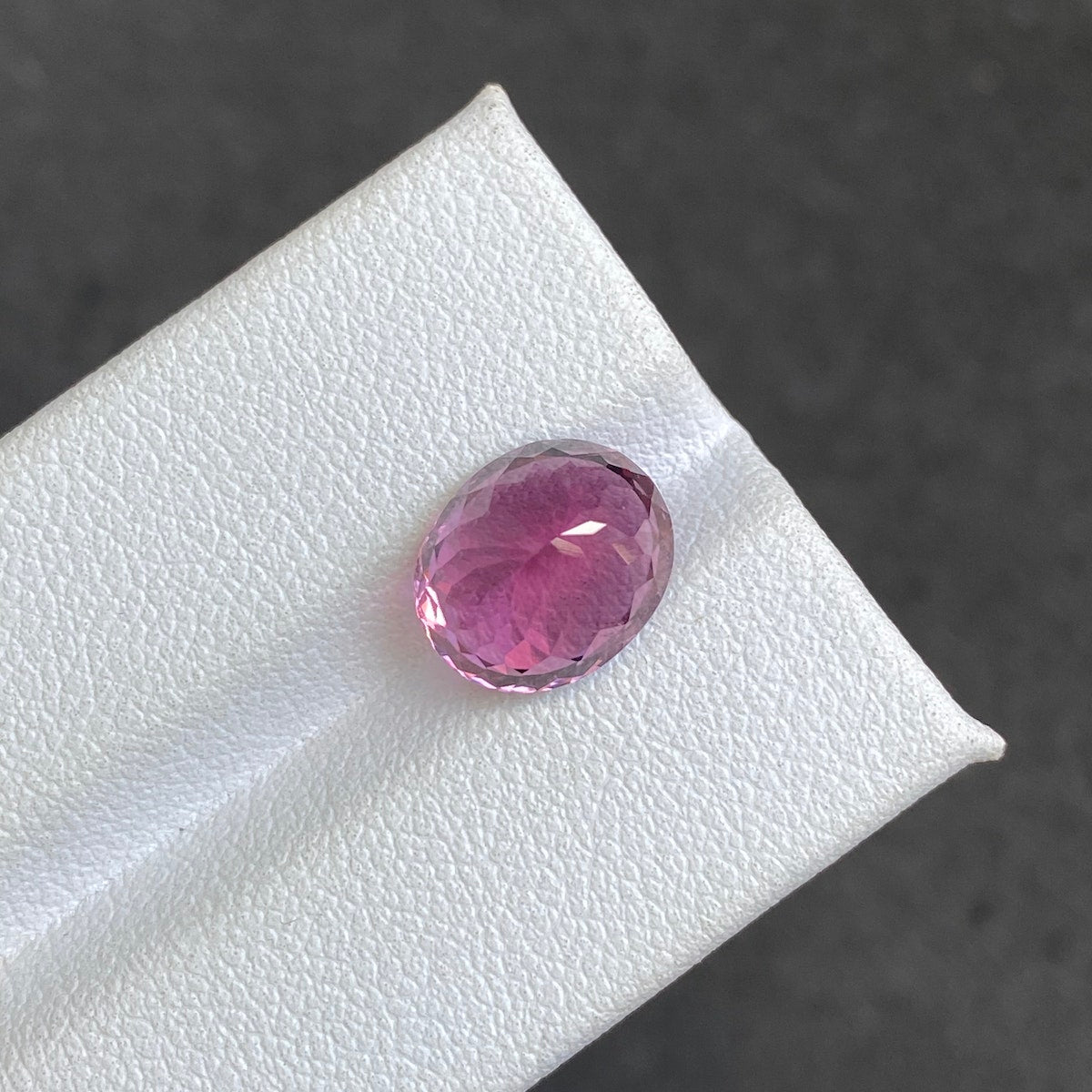 Une tourmaline rubellite ovale facettée de Bonnot Paris, pesant 4,35 carats, repose sur une surface blanche texturée sur un fond gris flou. Son aspect poli met en valeur une teinte rose vibrante qui reflète magnifiquement la lumière.