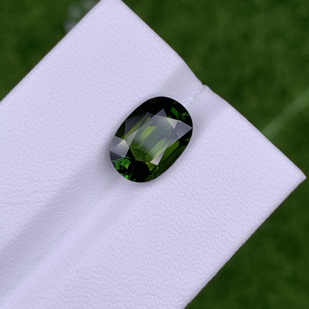 Gros plan de la pierre précieuse Tourmaline Chrome · 3,04 ct de Bonnot Paris reposant sur une surface blanche, mettant en valeur sa taille ovale et ses teintes vert foncé vibrantes avec des facettes reflétant la lumière, sur un fond vert flou rappelant l'herbe.