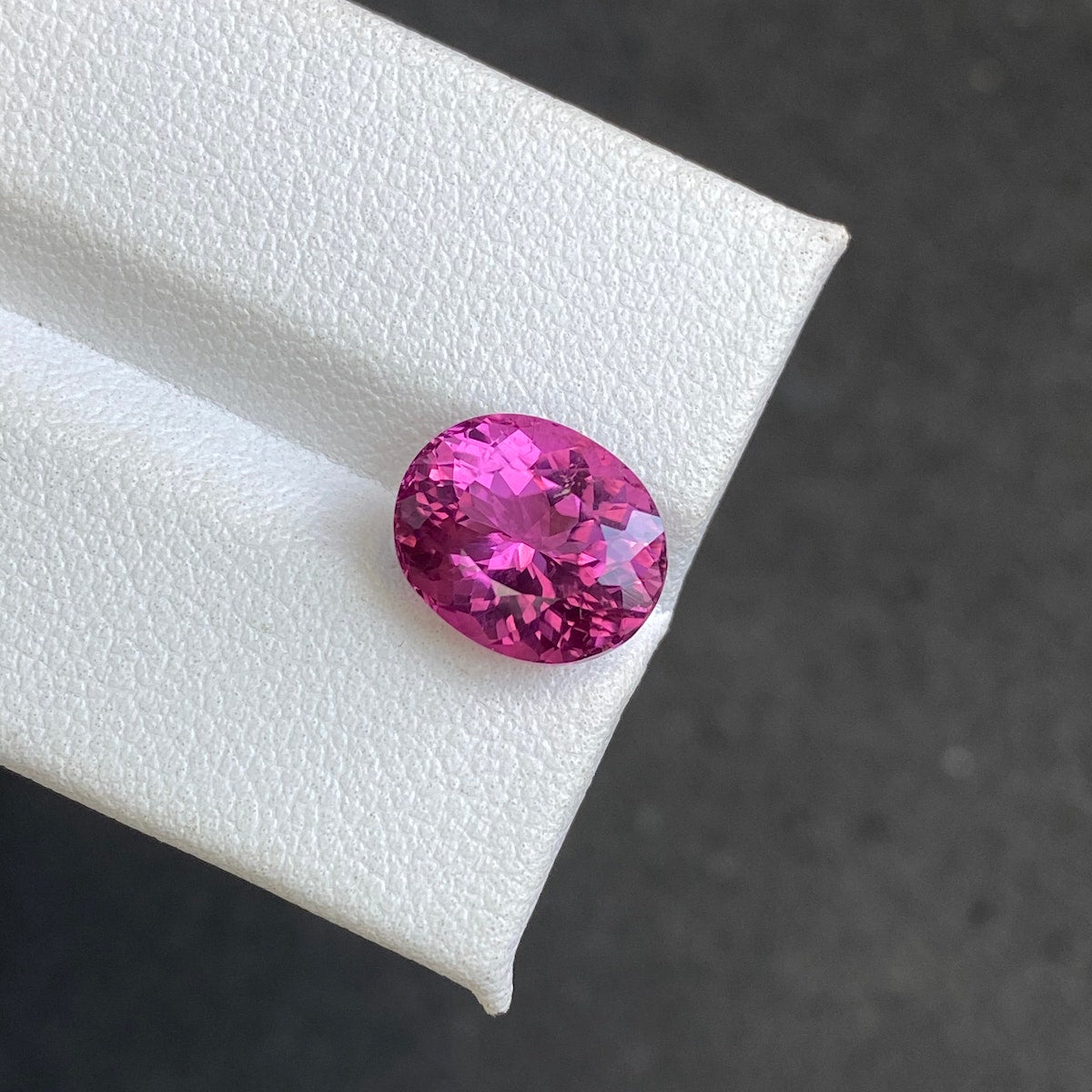 Une tourmaline rubellite ovale éclatante (4,35 carats) de Bonnot Paris repose sur une surface blanche texturée sur un fond sombre et flou. Ses facettes complexes et sa riche teinte scintillent de mille feux.