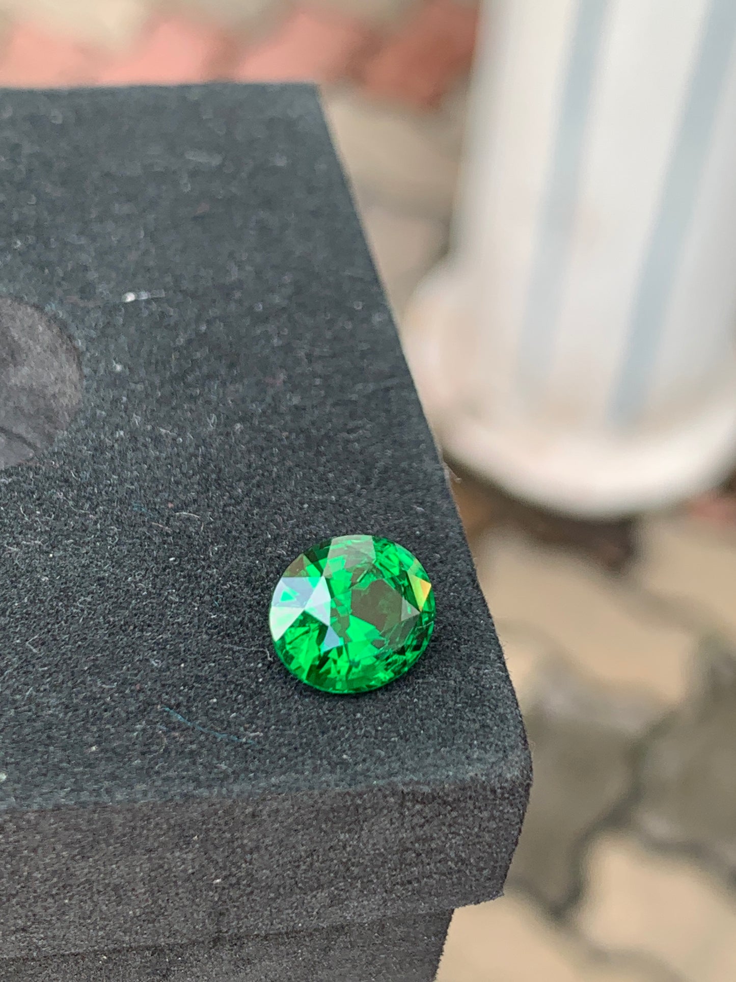 La tsavorite ronde facettée de 3,10 carats de Bonnot Paris repose sur une surface noire texturée, dont la teinte verte vibrante scintille de mille feux. Une colonne blanche et un pavé de briques en arrière-plan apportent une touche d'élégance inspirée de son origine tanzanienne.