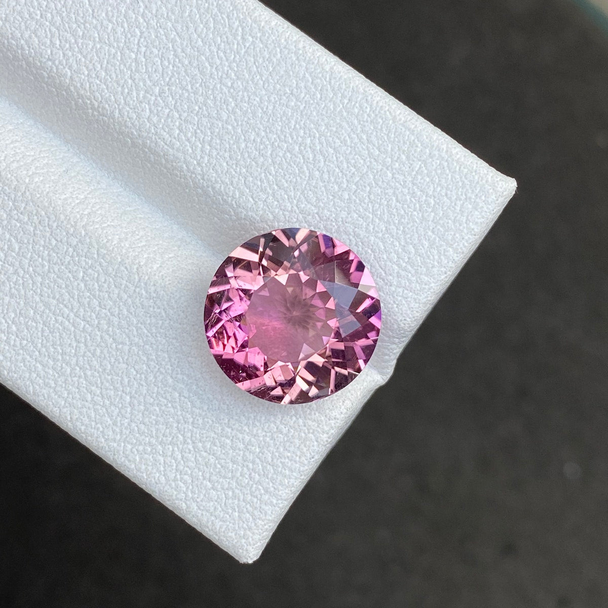 Une Tourmaline Cuprifère étincelante de Bonnot Paris, pesant 5,22 ct et présentant des facettes complexes, contraste brillamment sur une surface blanche texturée sur un fond sombre.