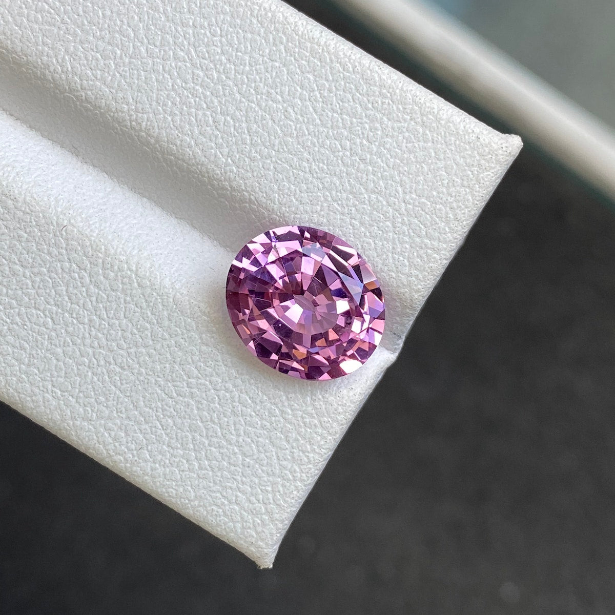 Un spinelle rose de 3,93 carats de Bonnot Paris repose sur une surface blanche texturée en angle, avec un fond sombre flou qui accentue sa couleur vibrante et ses facettes complexes.