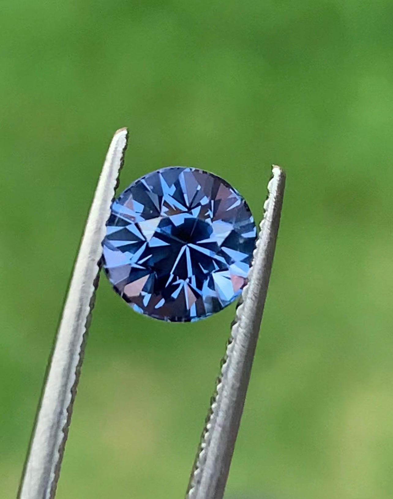 Une pierre précieuse ronde Spinelle Gris • 1,67 ct de Bonnot Paris est maintenue entre des pinces métalliques, sertie sur un fond vert flou, mettant en valeur sa brillance et son éclat facettés.