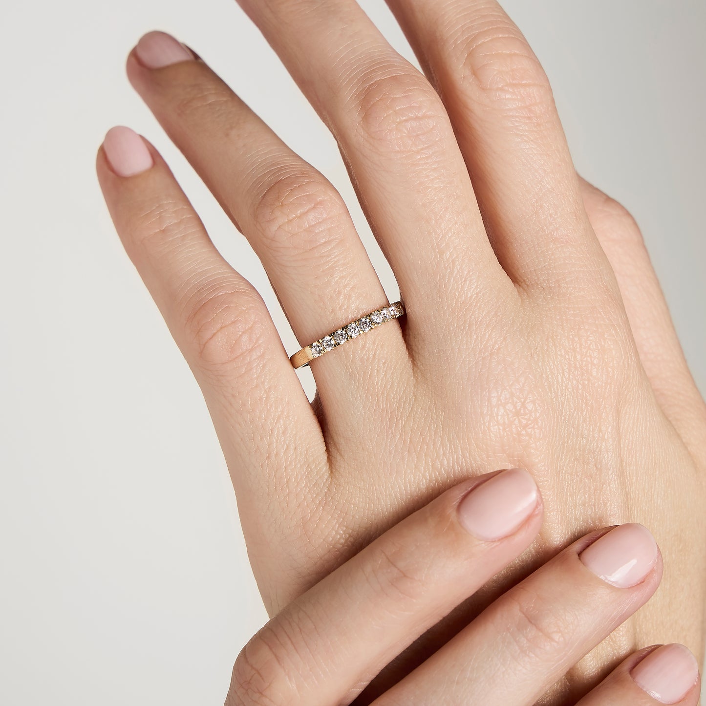 Un gros plan d'une main à la peau claire avec des ongles vernis rose pâle présente « Alliance La Discrète » de Bonnot Paris, une bande en or ornée de diamants naturels, sur un fond gris clair.