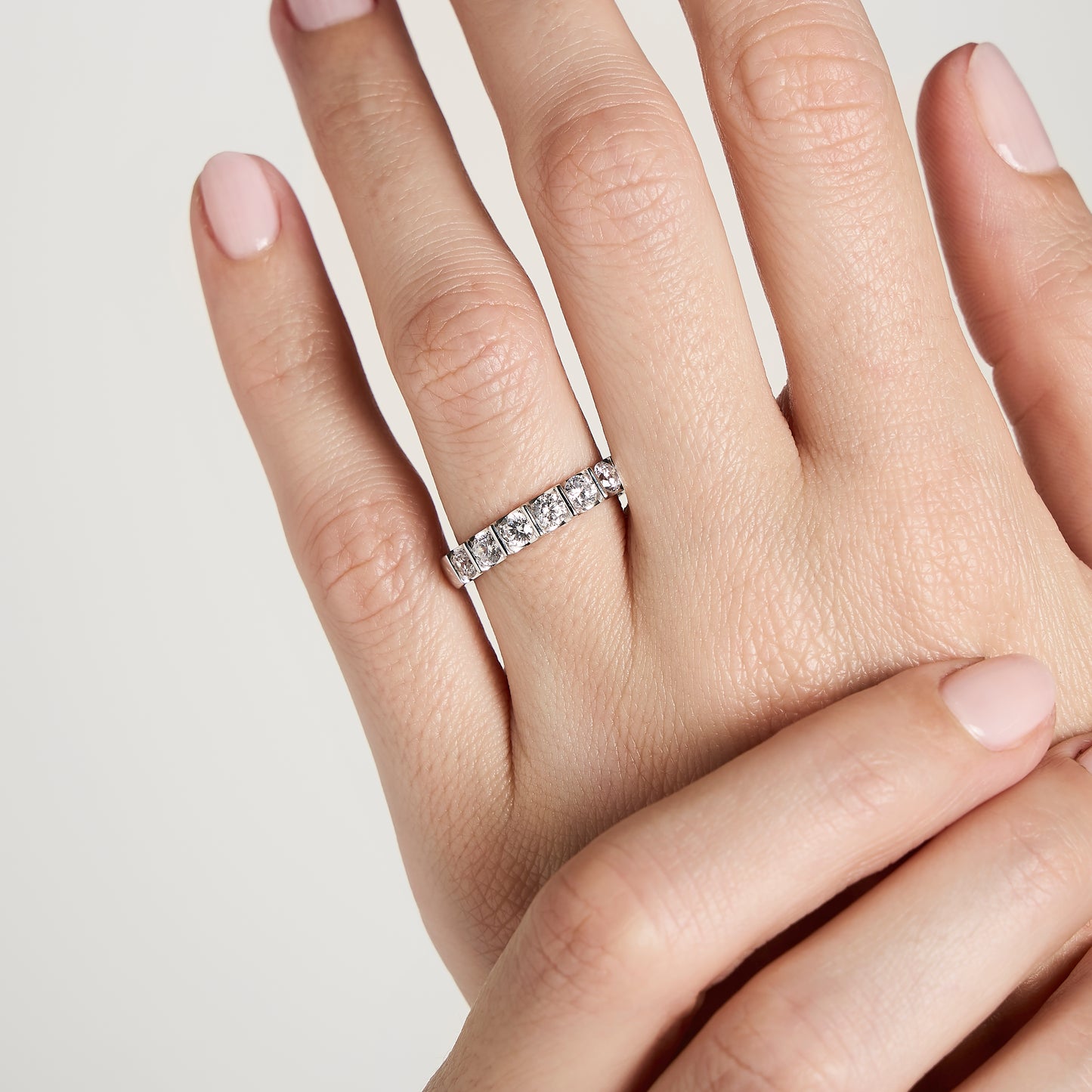 Une main aux ongles rose clair soigneusement manucurés arbore la bague en diamant Alliance La Subtile de Bonnot Paris à l'annulaire sur un fond blanc uni, tandis que l'autre main effleure délicatement la pierre taille rond-brillant, faisant partie d'une superbe collection de diamants naturels, soulignant son élégance.