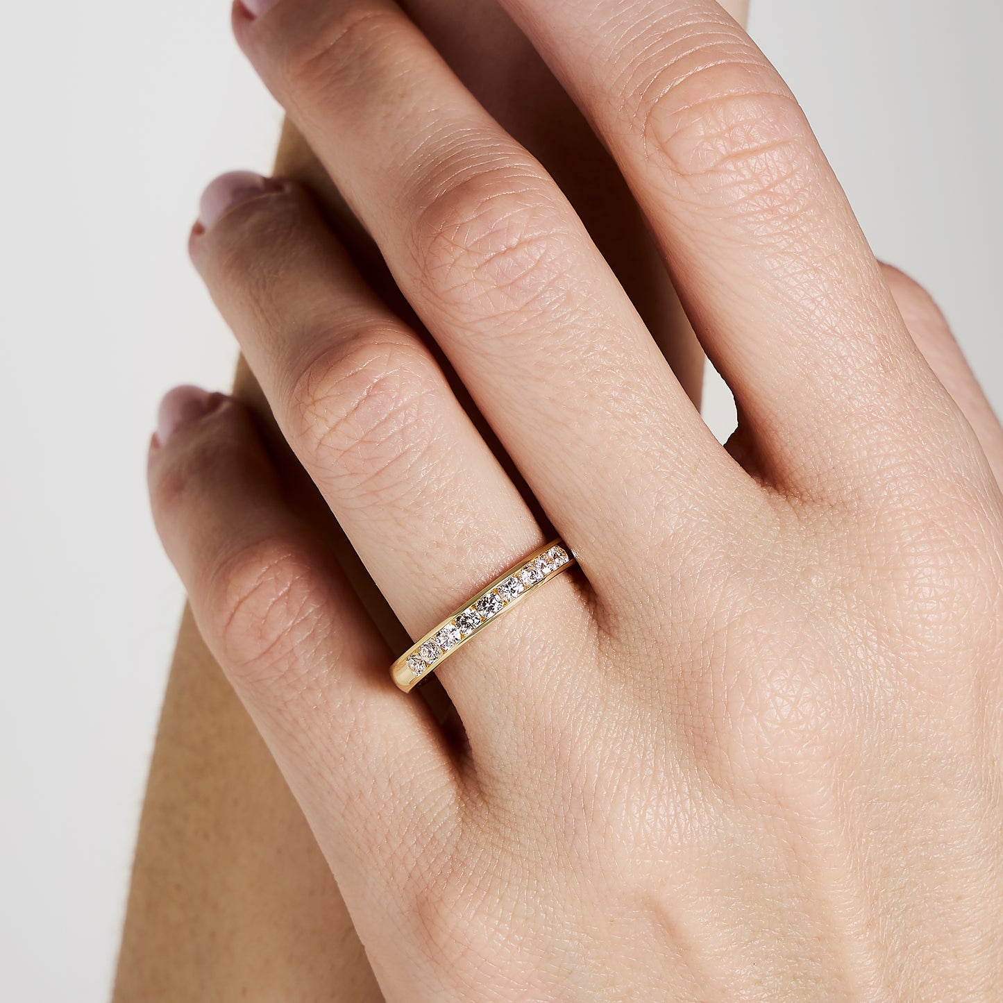 Une main ornée de l'Alliance La Délicate de Bonnot Paris, une magnifique bague en or sertie de minuscules diamants, à l'annulaire. La peau est claire et les ongles naturels arborent une teinte rosée. Le fond présente un doux dégradé de couleurs claires.