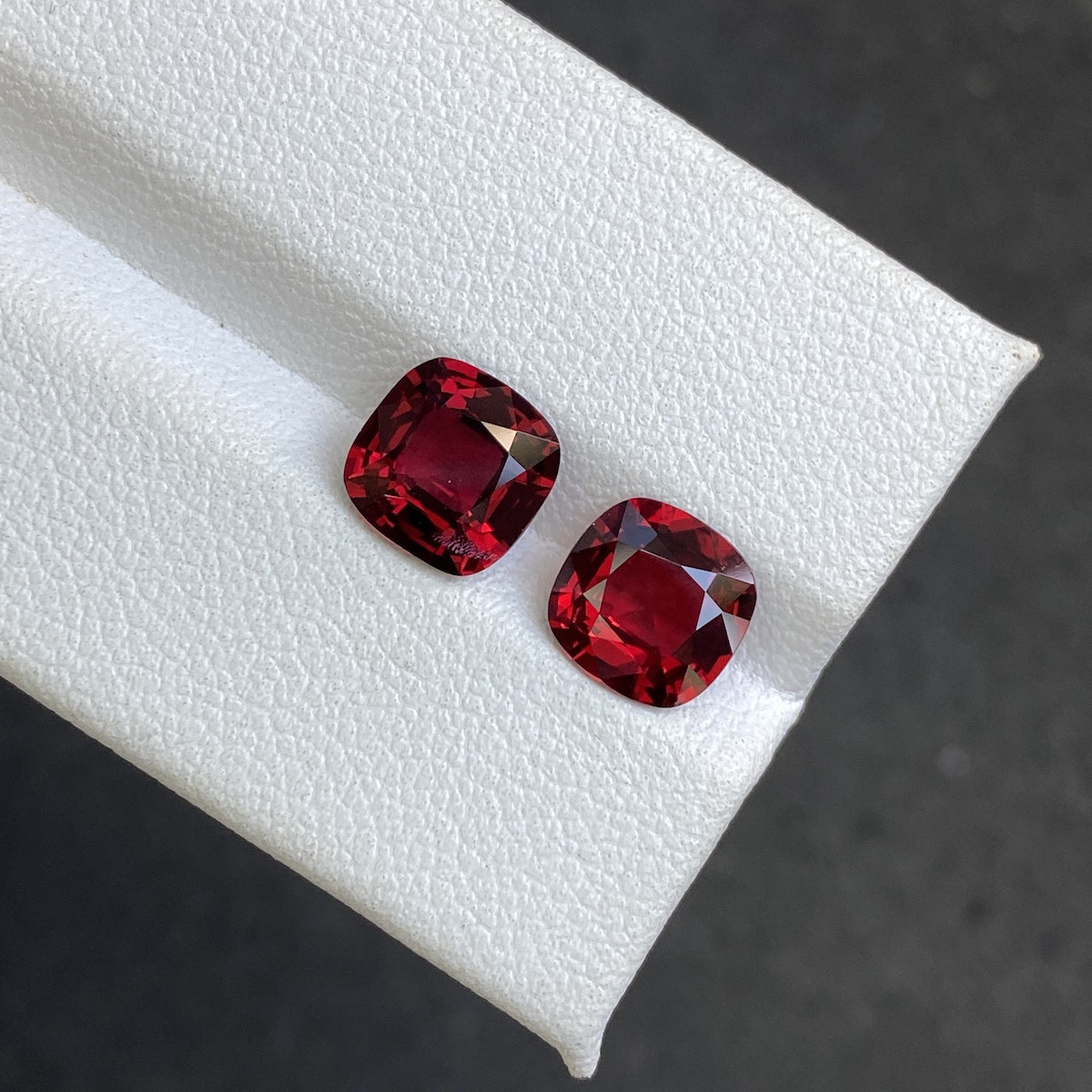 Deux pierres précieuses Bonnot Paris Spinelle Rouge de 3,49 carats, taille coussin, sont mises en valeur sur une surface blanche texturée, d'un rouge profond et vibrant. Elles se juxtaposent sur un fond gris foncé, accentuant leur éclat.