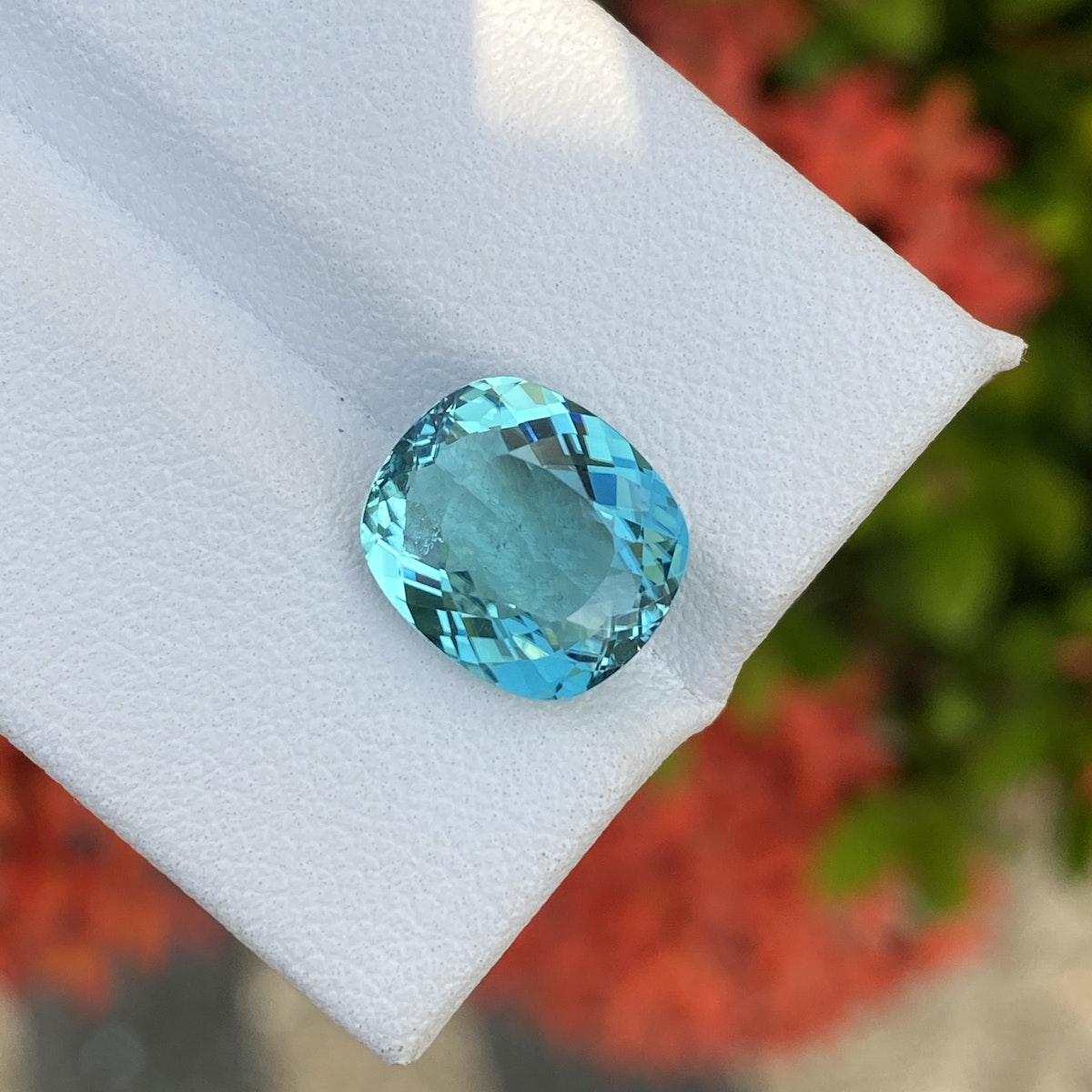 Une tourmaline paraïba taille coussin de 4,08 carats de Bonnot Paris repose sur une surface blanche, mettant en valeur sa couleur bleu vif et ses facettes complexes. Un fond rouge et vert flou ajoute du contraste.
