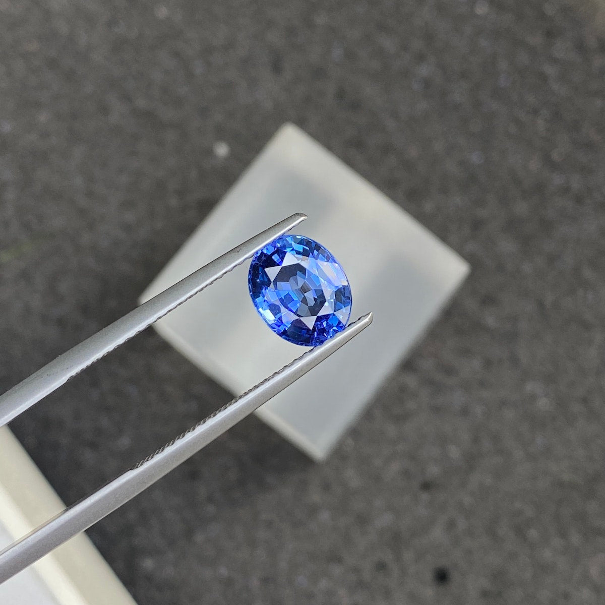 Gros plan du Saphir Bleu · 2,04ct de Bonnot Paris, une pierre précieuse ovale bleue taillée en facettes maintenue par une pince à épiler sur un fond flou, mettant en valeur sa clarté et sa couleur vibrante.