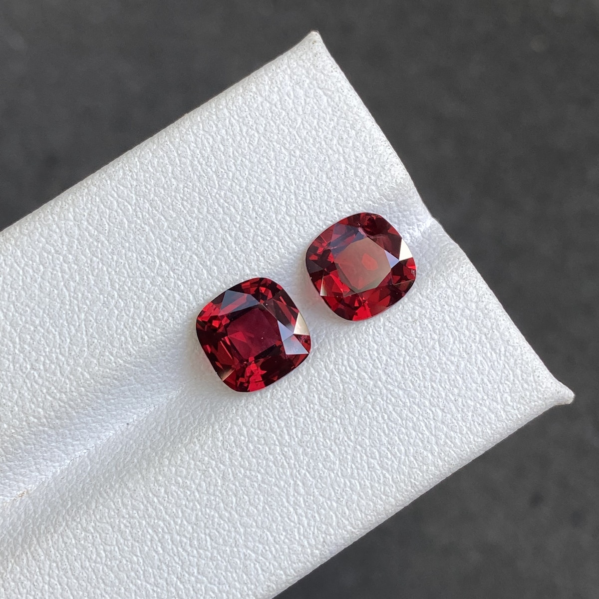 Deux spinelles rouges (3,49 carats) de Bonnot Paris, taillées en carré et dotées d'une finition brillante d'un rouge profond, reposent sur une surface blanche texturée. Un fond gris foncé flou rehausse leur teinte vive.