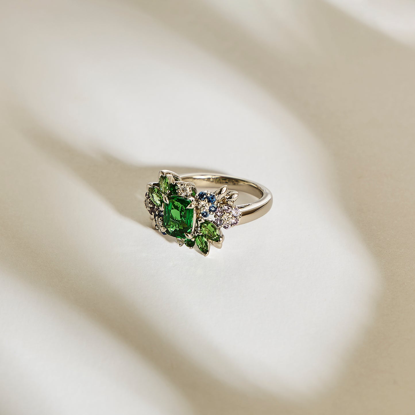 La bague en or Green Bloom - Tsavorite de Bonnot Paris présente une grande pierre précieuse verte entourée de plus petites tsavorites Grenat et de pierres claires, élégamment présentées sur une surface légèrement texturée et de couleur claire avec des ombres douces.