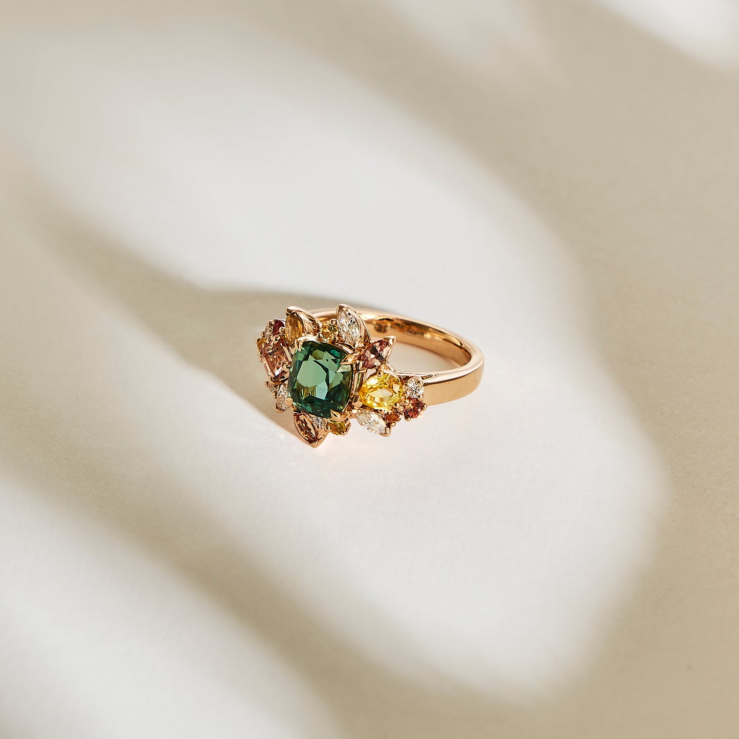 Le Sea Bloom - Tourmaline de Bonnot Paris présente une pierre précieuse verte centrale, probablement de la tourmaline bleu-vert, entourée de pierres multicolores plus petites dans un motif floral, reposant sur une surface doucement éclairée projetant de douces ombres.