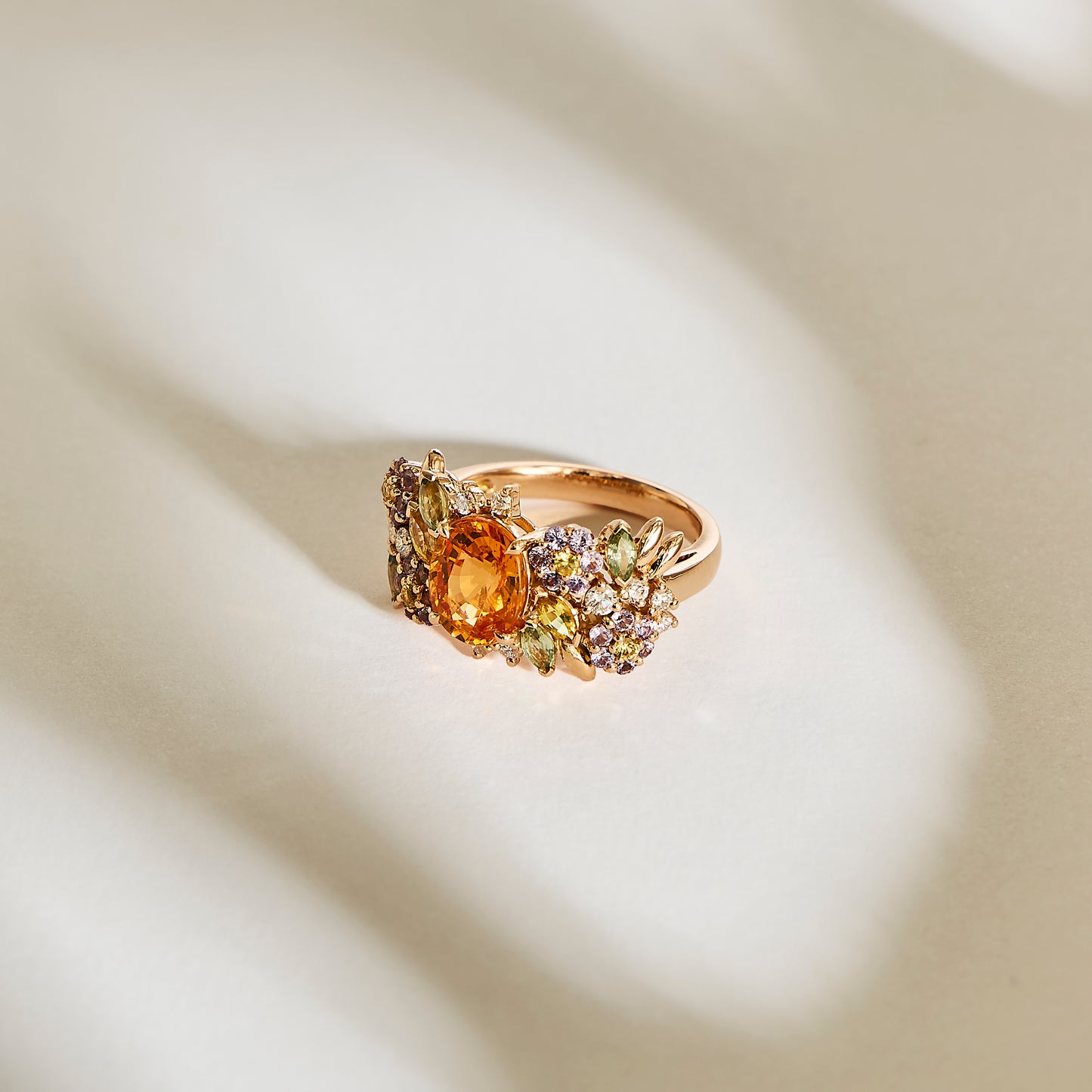La bague Golden Bloom - Grenat Mandarin de Bonnot Paris présente une spessartite grenat centrale entourée d'une fleur de couleur de pierres violettes et vertes plus petites dans un design floral exquis, le tout reposant sur une surface texturée doucement éclairée.