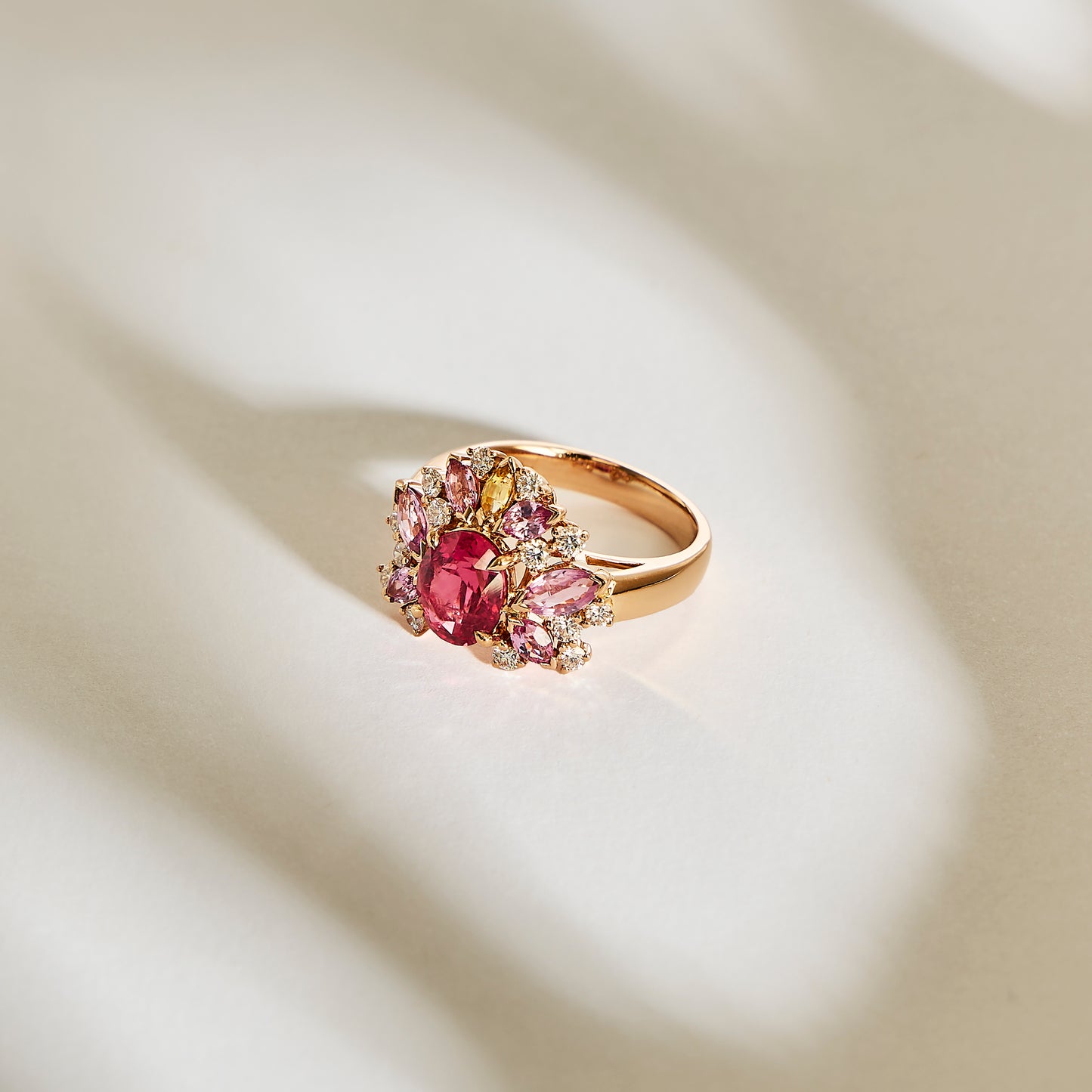 La bague en or Bonnot Paris "Rose Bloom - Spinelle Mahenge" présente une spinelle Mahenge centrale proéminente, entourée de pierres roses et claires plus petites, reposant élégamment sur une surface doucement éclairée et légère.