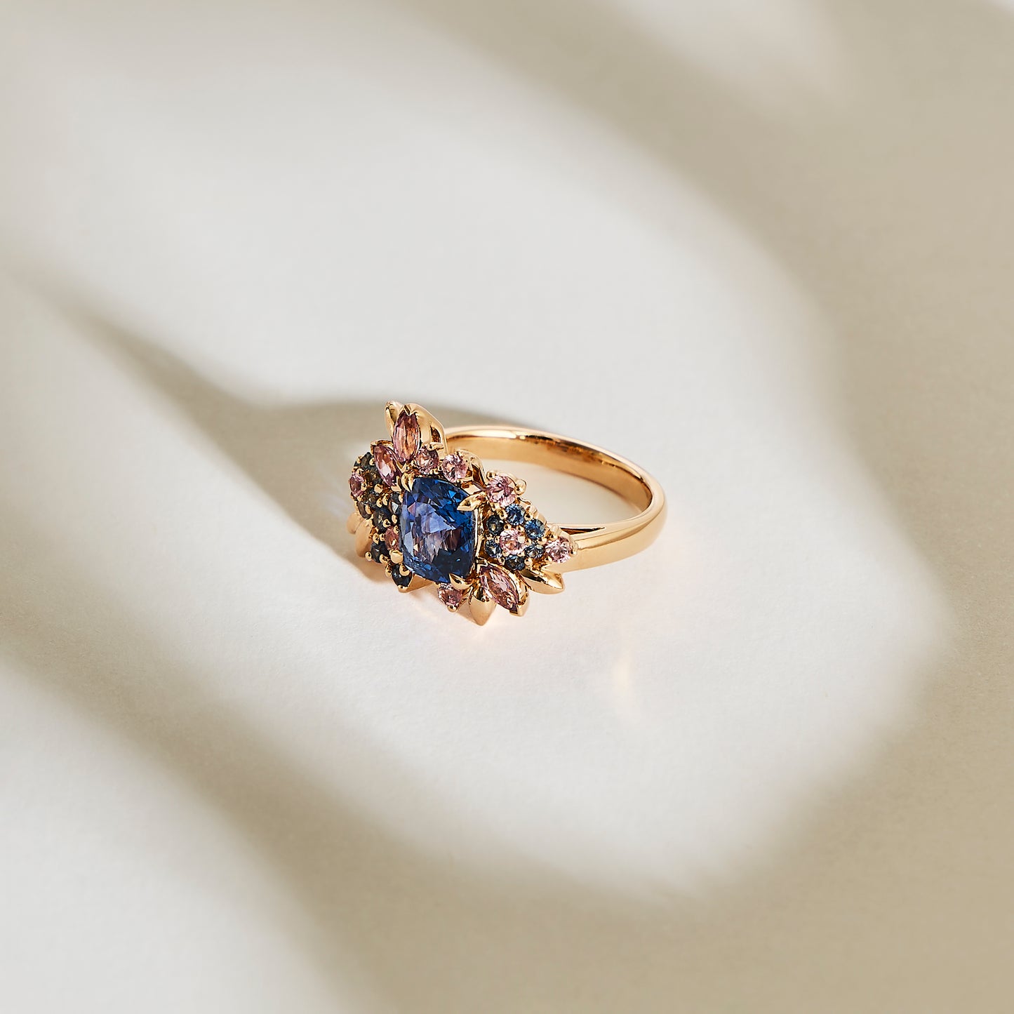 La bague Navy Bloom - Saphir de Bonnot Paris présente un grand saphir bleu ovale, entouré de petites pierres précieuses colorées, projetant une ombre sur une surface texturée doucement éclairée.