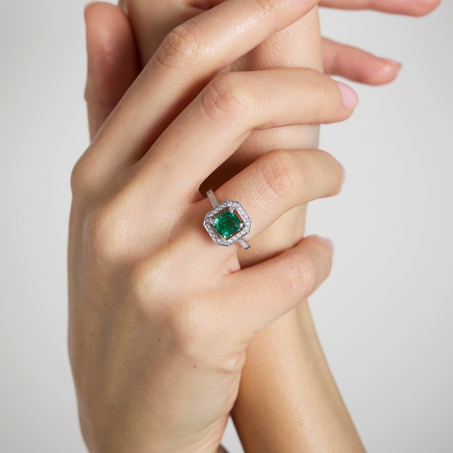 Gros plan sur une main portant la Bague Émeraude • 1,22 carat de Bonnot Paris. Sa pierre zambienne taille émeraude est entourée d'un halo de diamants sertis dans un élégant métal blanc, exprimant un style Art déco. La main au poli neutre en saisit une autre sur un fond sobre.