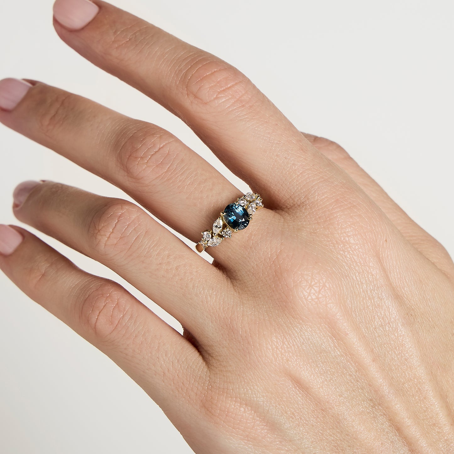 Une main aux ongles rose clair manucurés arbore la Bague Saphir Teal de Bonnot Paris • 1,14 ct, ornée d'un saphir bleu-vert entouré de diamants blancs sur une élégante bande argentée, le tout sur fond blanc.