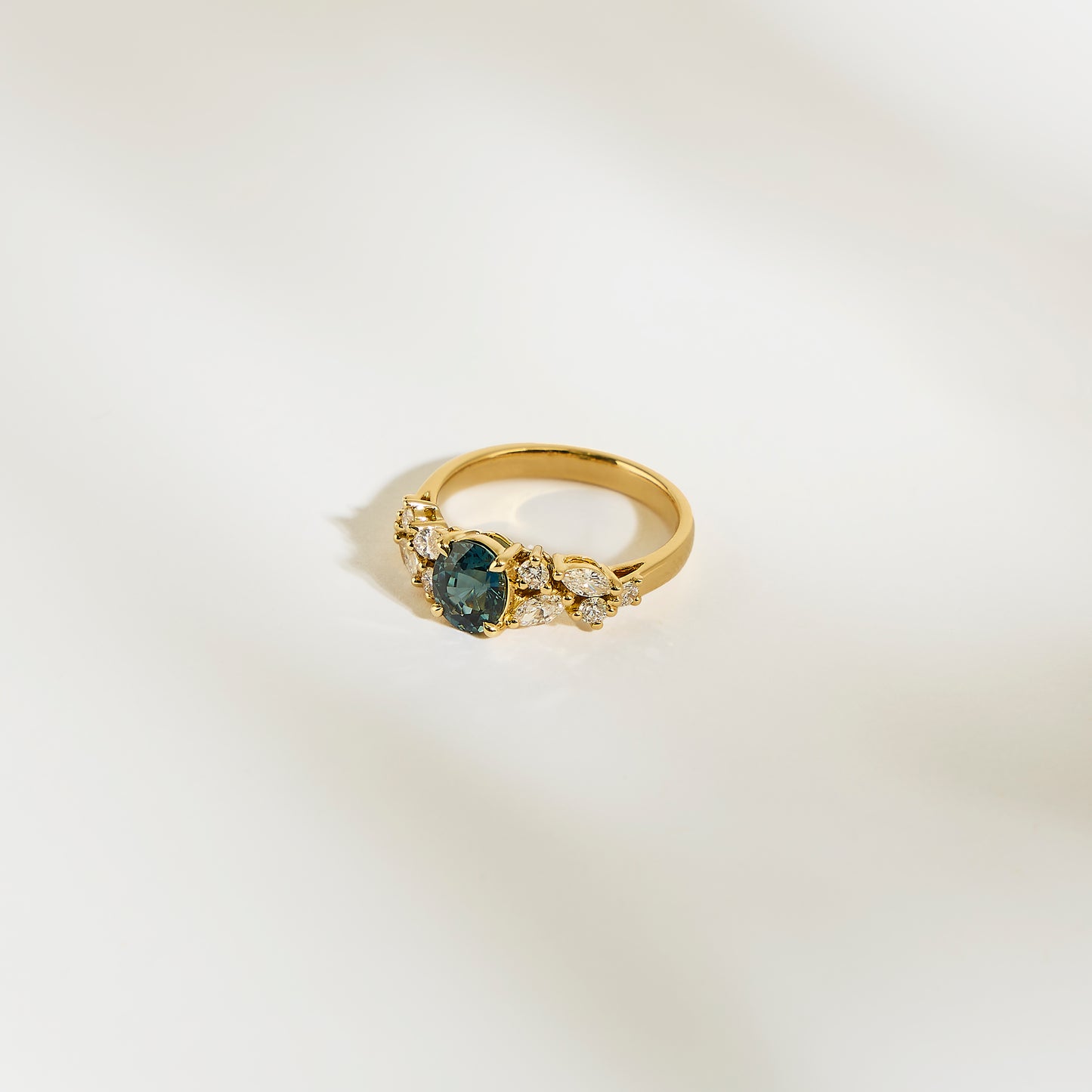 La Bague Saphir Teal de Bonnot Paris • 1,14 carat est ornée d'un saphir bleu sarcelle central entouré de petits diamants blancs. Sur un fond crème clair, ses détails raffinés et son design élégant en font une bague de fiançailles idéale.