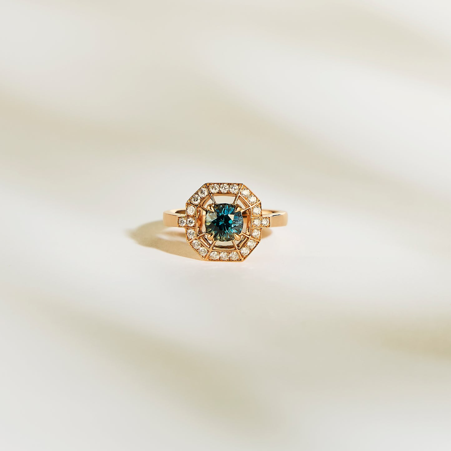 La Bague Saphir Teal de Bonnot Paris • 1,16 ct est une superbe bague de fiançailles en or avec un saphir sarcelle central et un halo de diamants blancs étincelants, présentés sur une surface douce et claire.