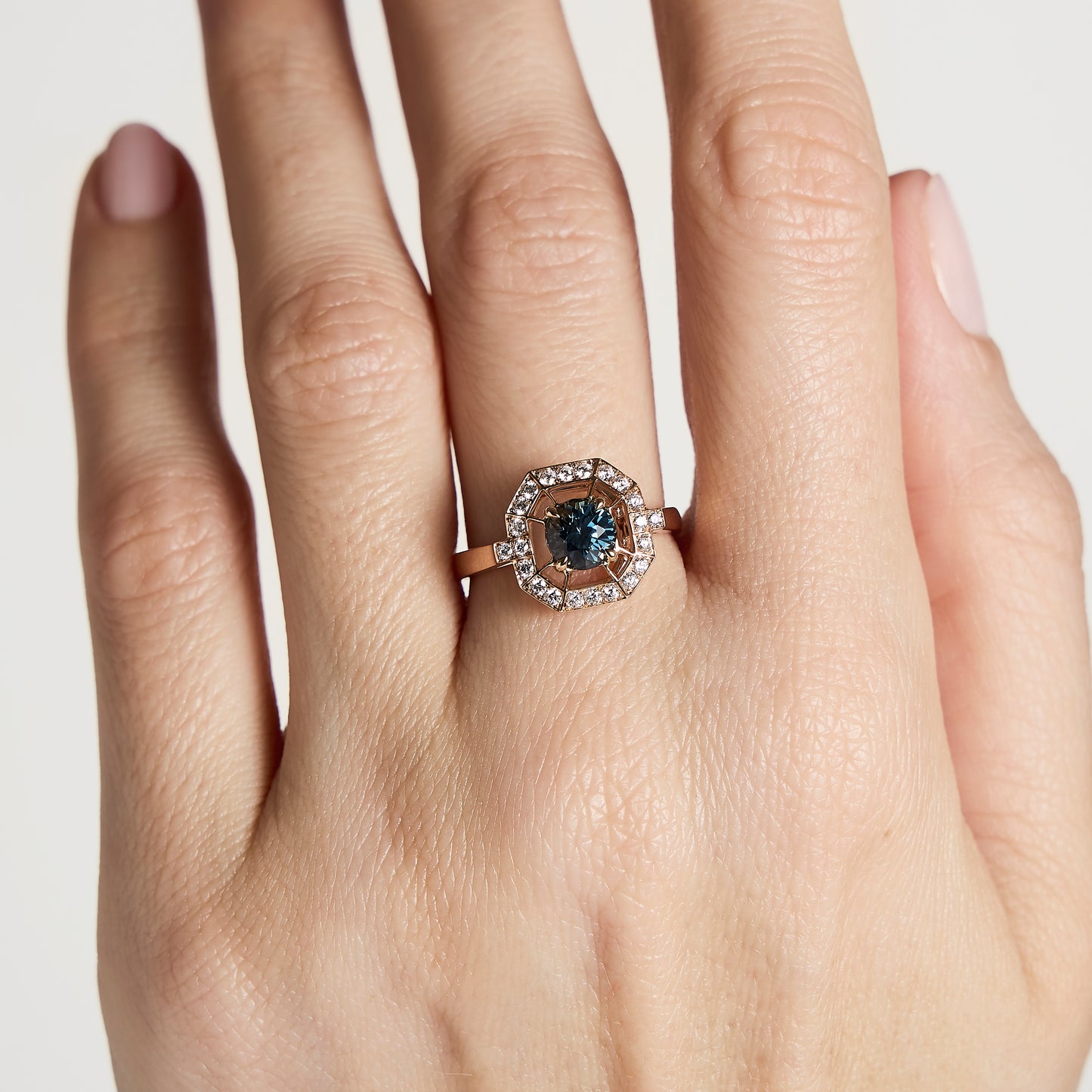 Une main avec un ongle soigneusement manucuré porte la bague Bague Saphir Teal de Bonnot Paris • en or 1,16 carat, mettant en valeur un saphir sarcelle central entouré d'un halo de diamants blancs étincelants sur un fond clair neutre.