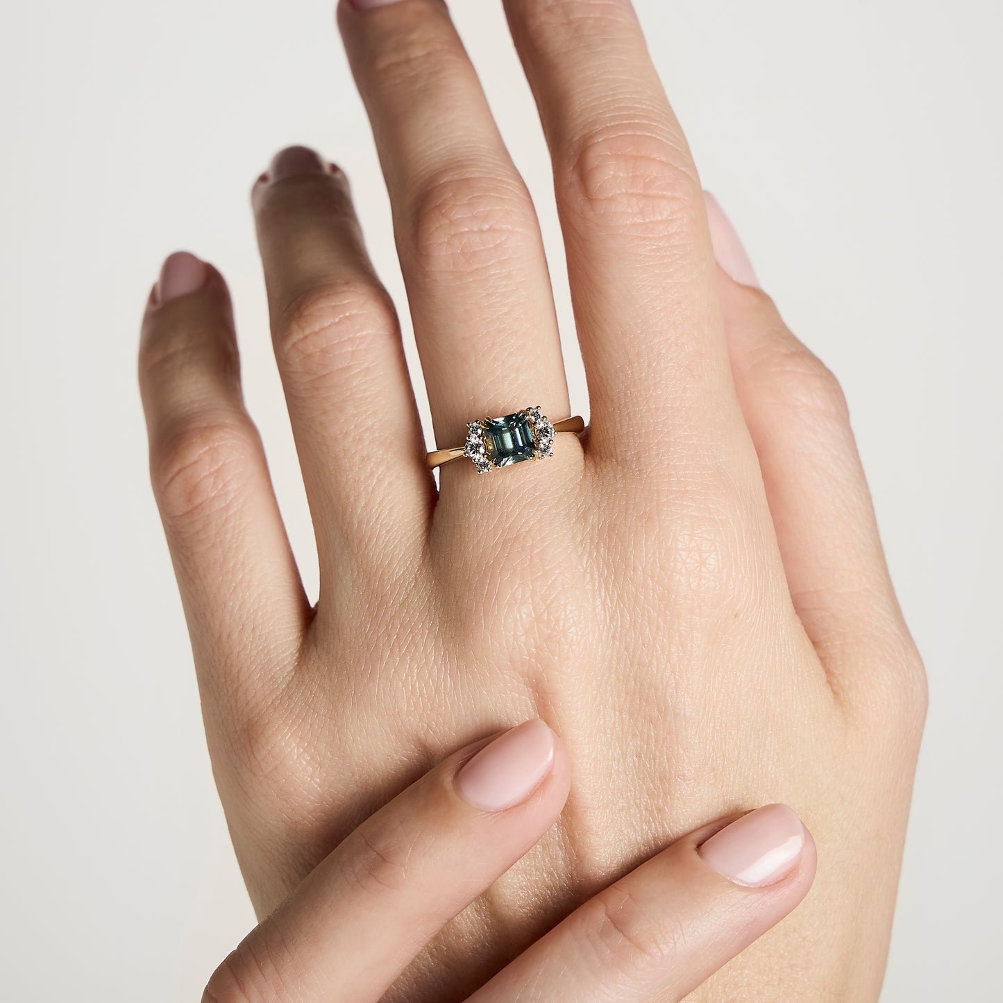 Une main aux ongles roses parfaitement manucurés arbore l'exquise Bague Saphir Teal de Bonnot Paris • 1,24 ct, avec un captivant saphir sarcelle taille émeraude serti en or, orné de délicats diamants blancs sur un fond clair - idéale comme bague de fiançailles pour quelqu'un de spécial.