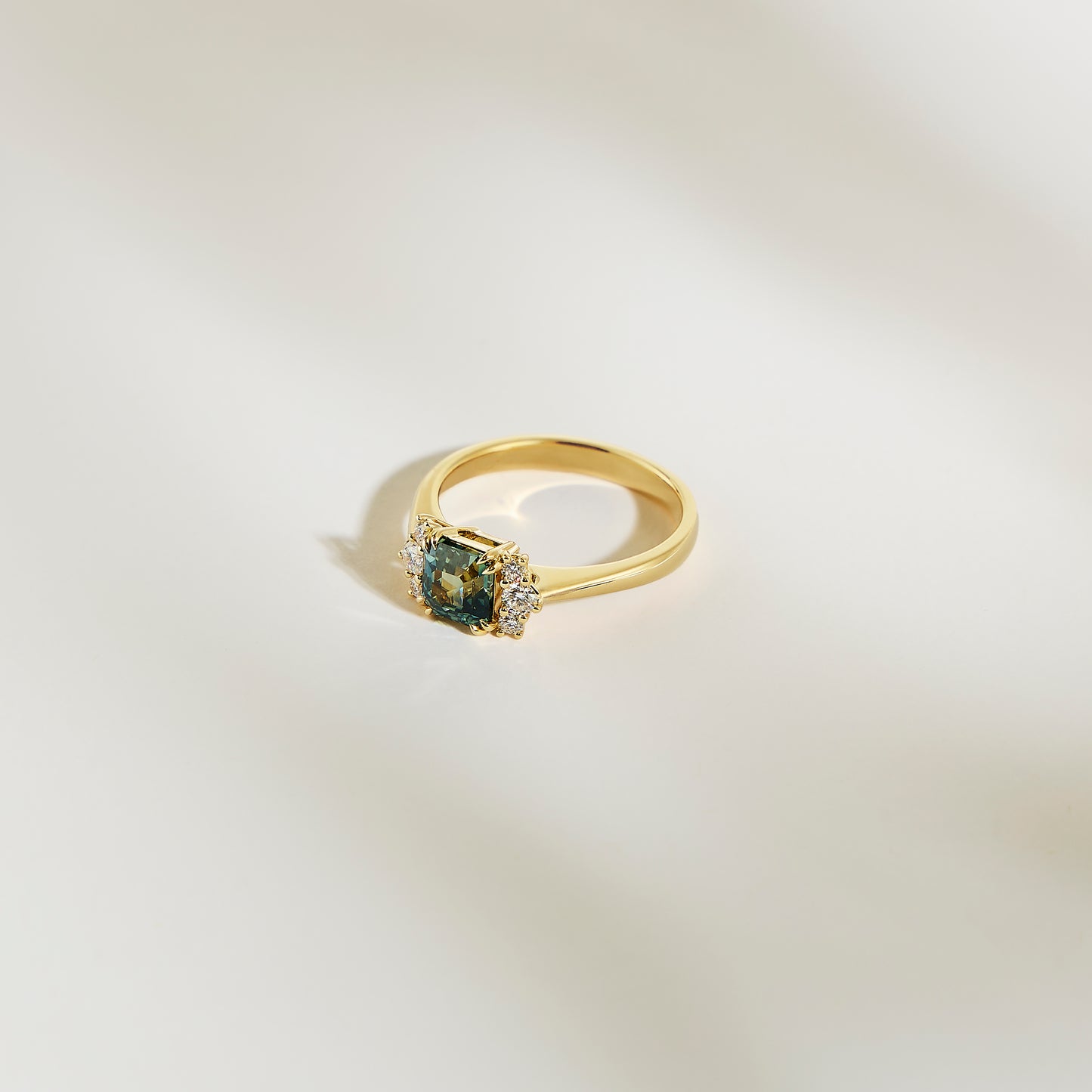 La Bague Saphir Teal • 1,24ct de Bonnot Paris, une bague en or avec un saphir sarcelle taille carrée en son centre et entouré de petits diamants blancs, est présentée sur une surface lisse de couleur claire.