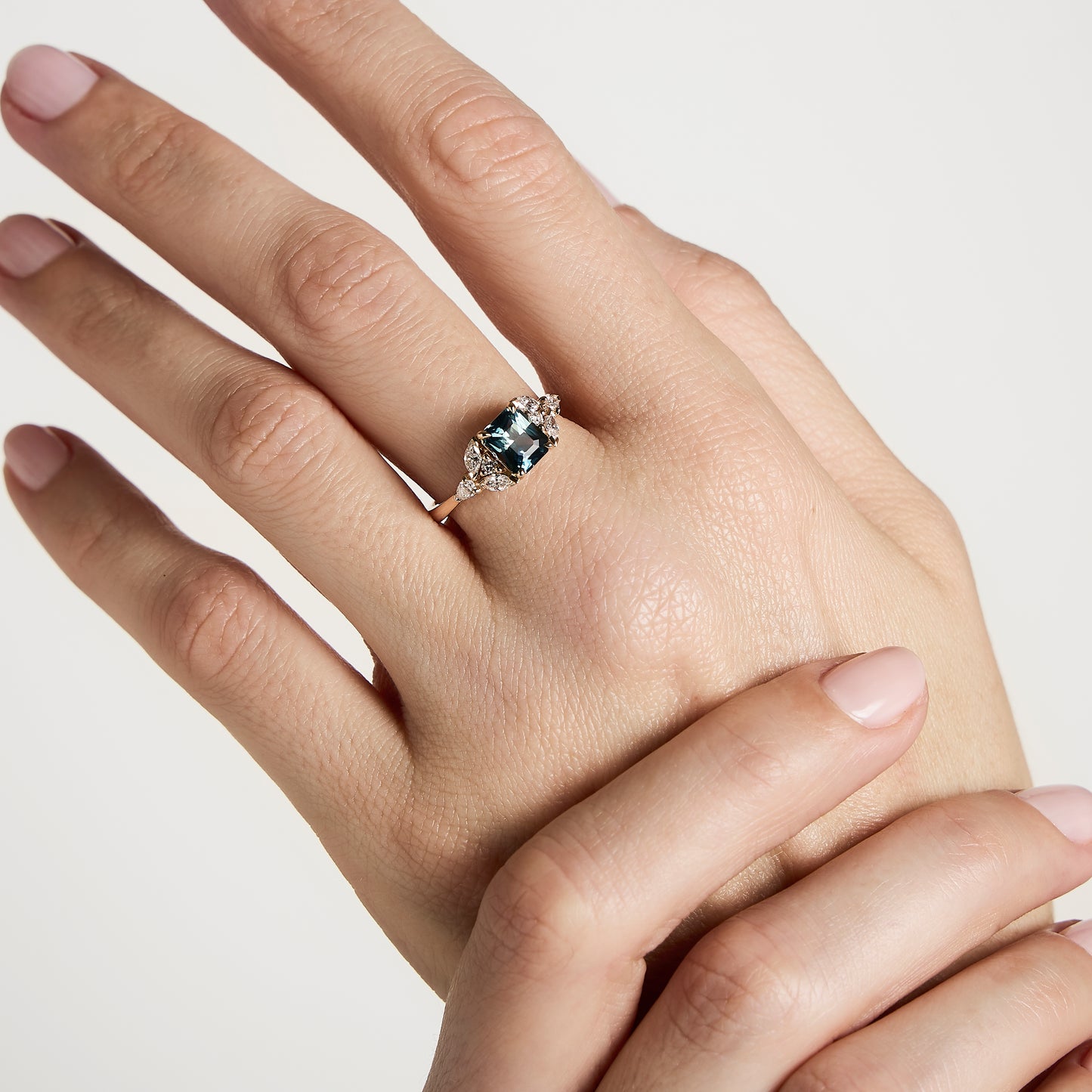 Une main aux ongles manucurés arbore une Bague Saphir Teal Bonnot Paris de 1,56 carat. La bague est ornée d'un grand saphir rectangulaire turquoise encerclé de petits diamants blancs. Les doigts, élégamment posés, se détachent sur un fond blanc uni, mettant en valeur cette magnifique bague de fiançailles.