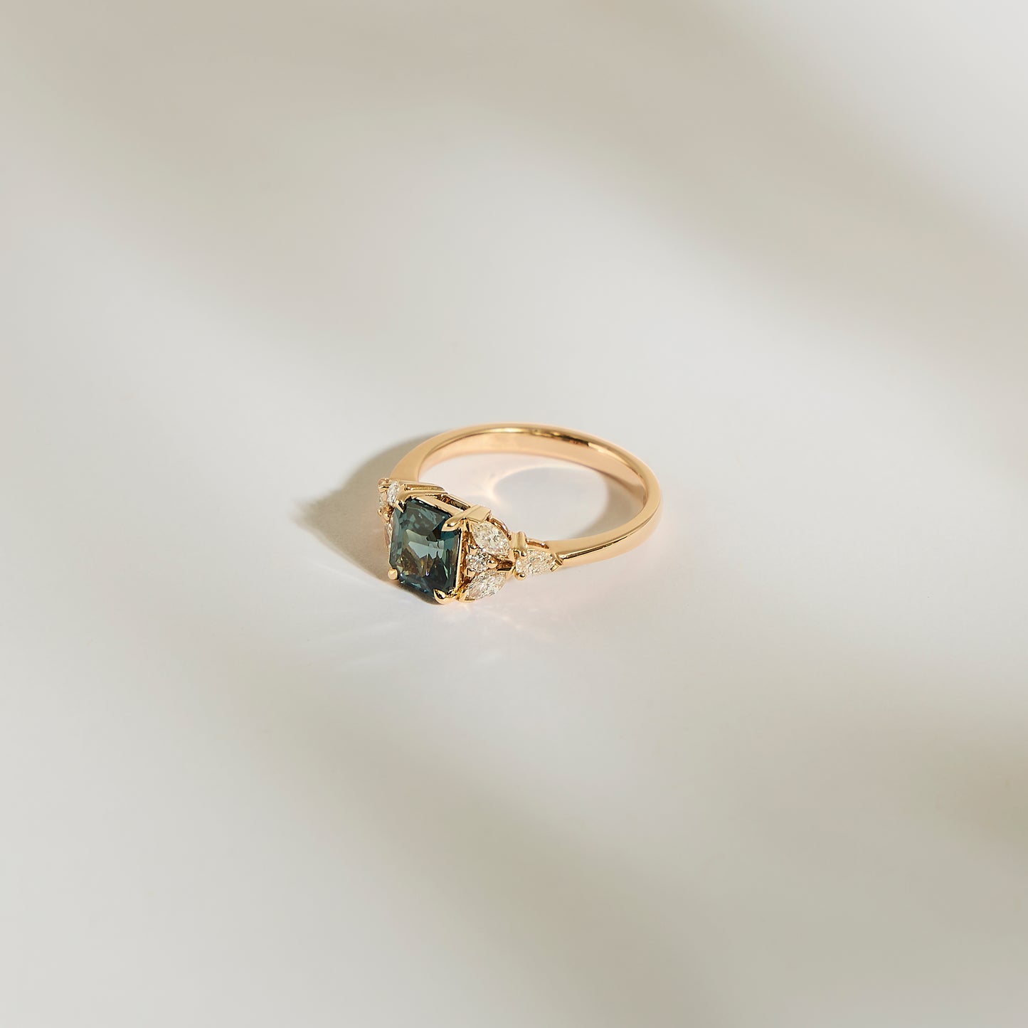 La Bague Saphir Teal • 1,56ct de Bonnot Paris est une bague en or présentant un grand centre rectangulaire en pierre précieuse noire, entouré de petits diamants blancs sur une surface lisse et légère, projetant une ombre douce.