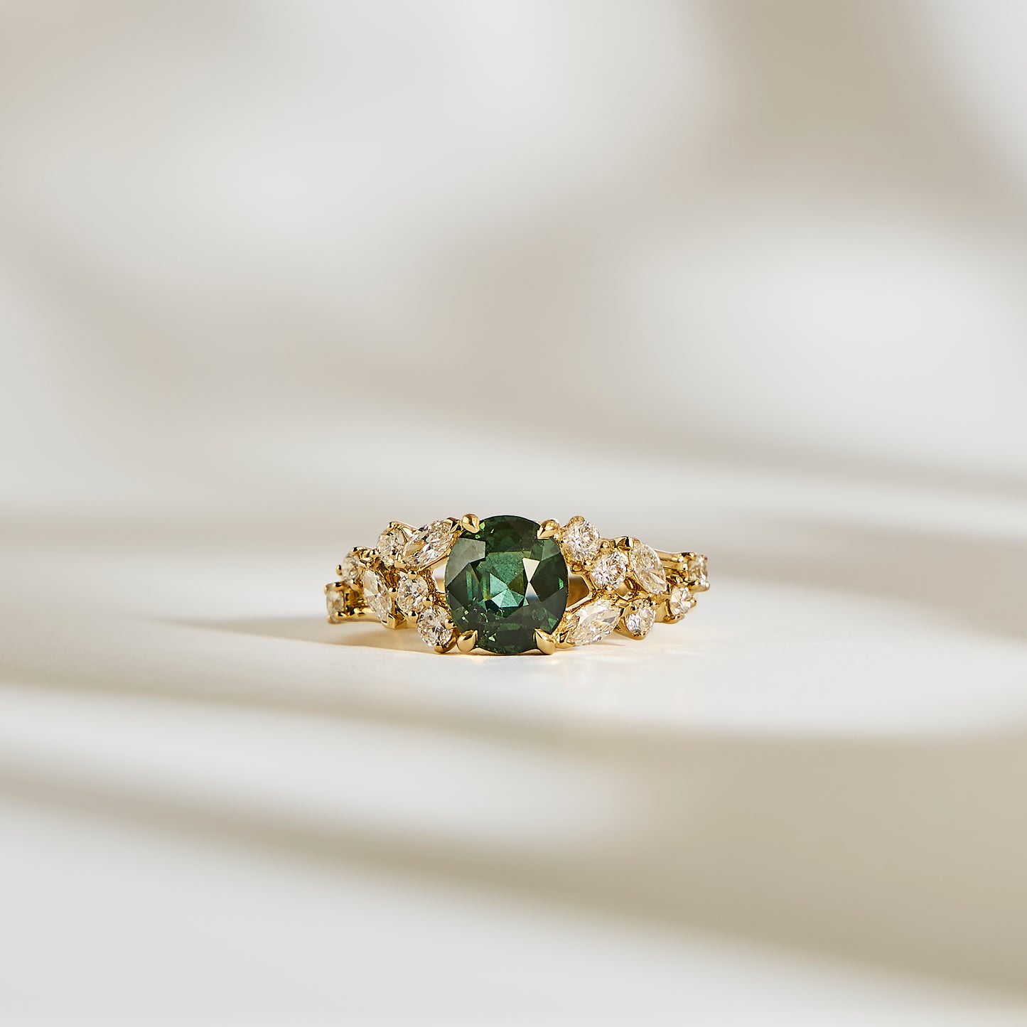 La Bague Saphir Teal • 2,07ct de Bonnot Paris, une bague en or avec un centre en saphir bleu sarcelle saisissant entouré de pierres claires, repose élégamment sur une surface crème doucement éclairée avec des ombres douces.