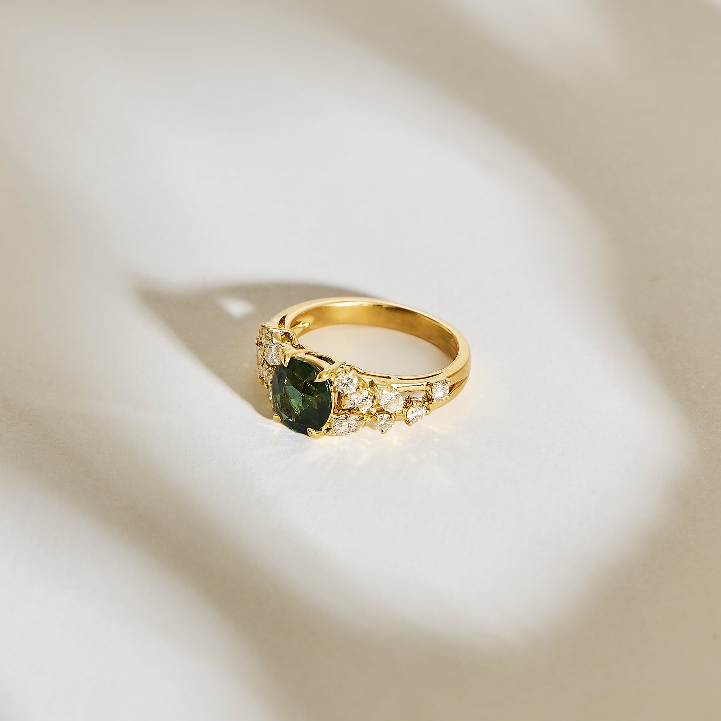 La Bague Saphir Teal • 2,07ct de Bonnot Paris présente une pierre précieuse verte en forme de cœur bordée de petites pierres claires, magnifiquement sertie sur un fond neutre doucement éclairé par des ombres douces.