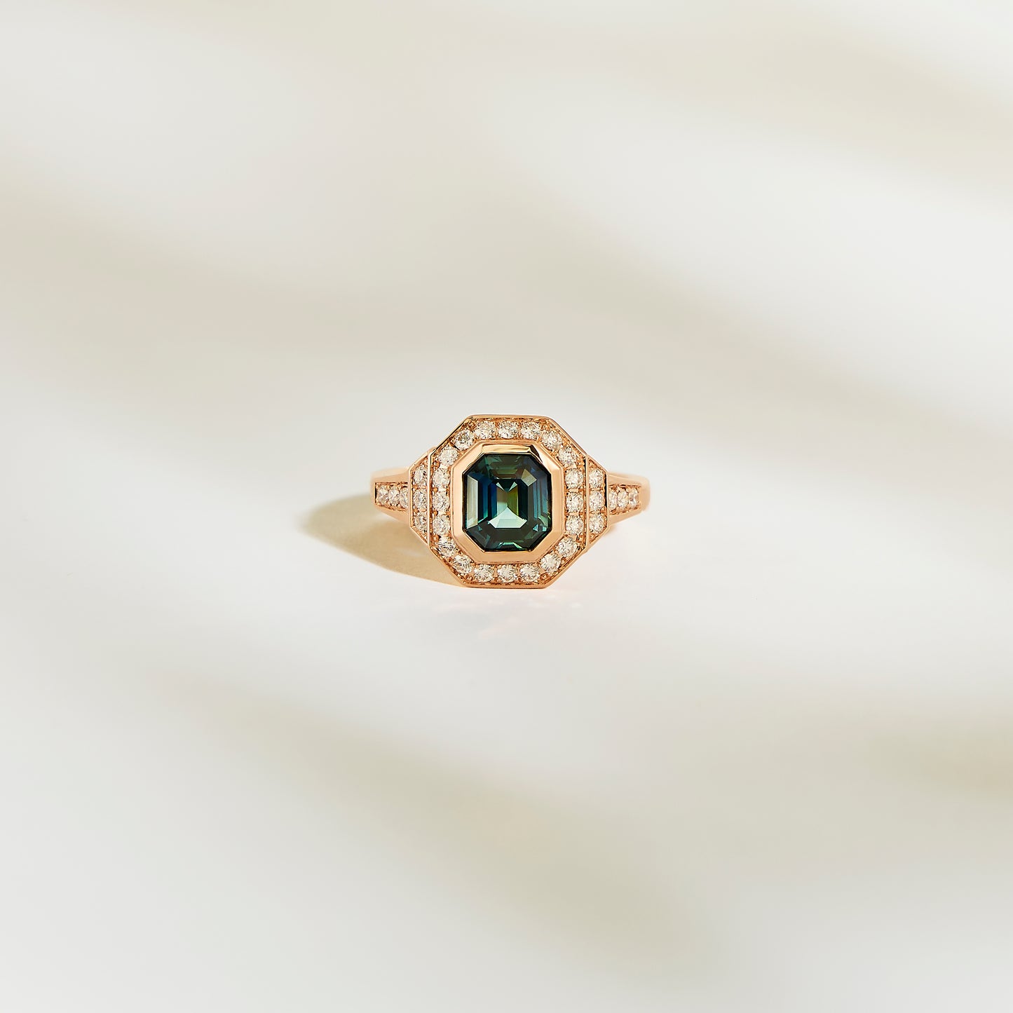 La Bague Saphir Teal de Bonnot Paris, une bague en or ornée d'une pierre précieuse vert foncé taille émeraude de 1,56 ct entourée d'un halo de petits diamants blancs, est élégamment mise en valeur sur un fond doux et clair.